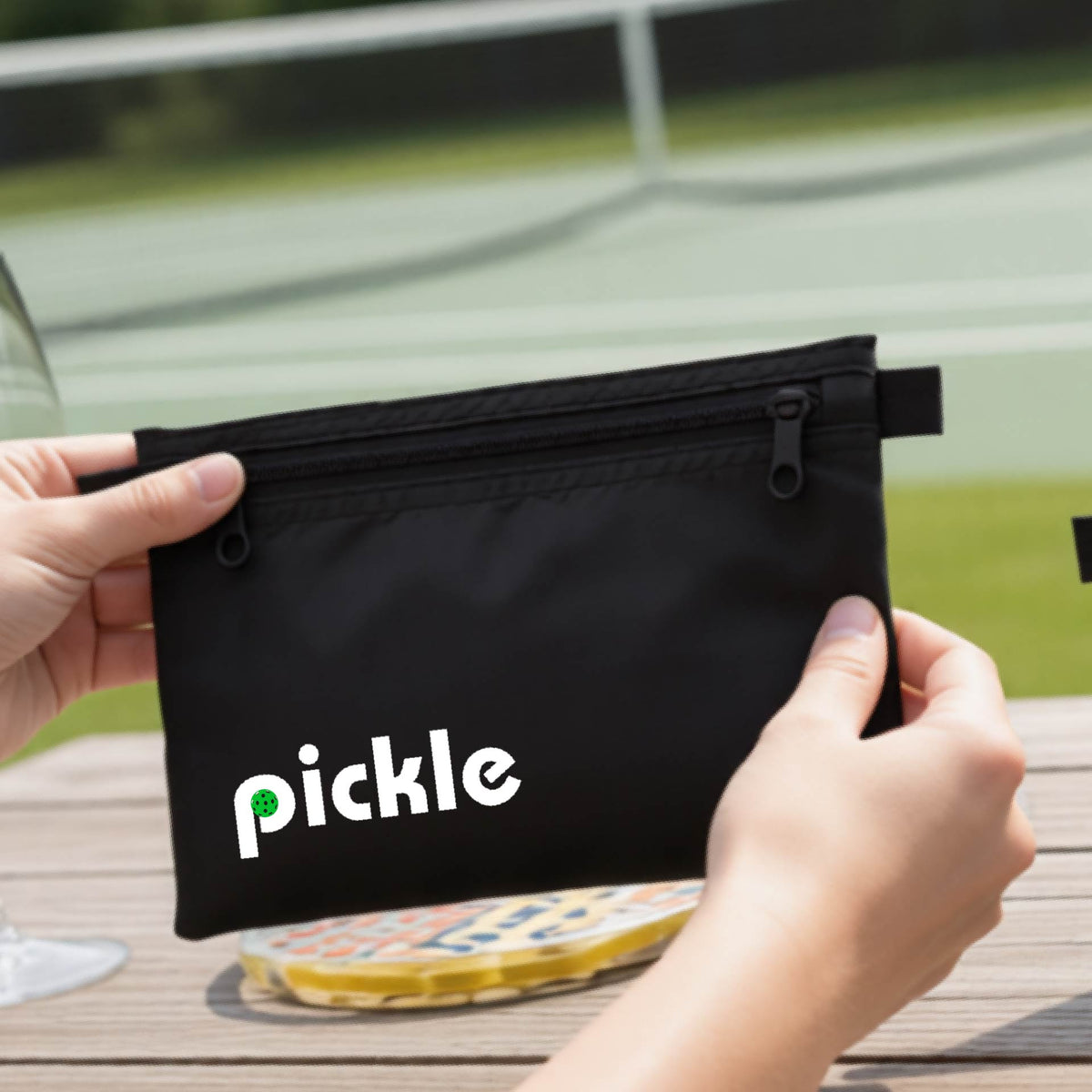 A close-up of a black pickleball polyester cosmetic bag with a white graphic of ‘PICKLE’ with a green pickleball integrated into the letter P printed on the upper right corner, in front of a pickleball court. 