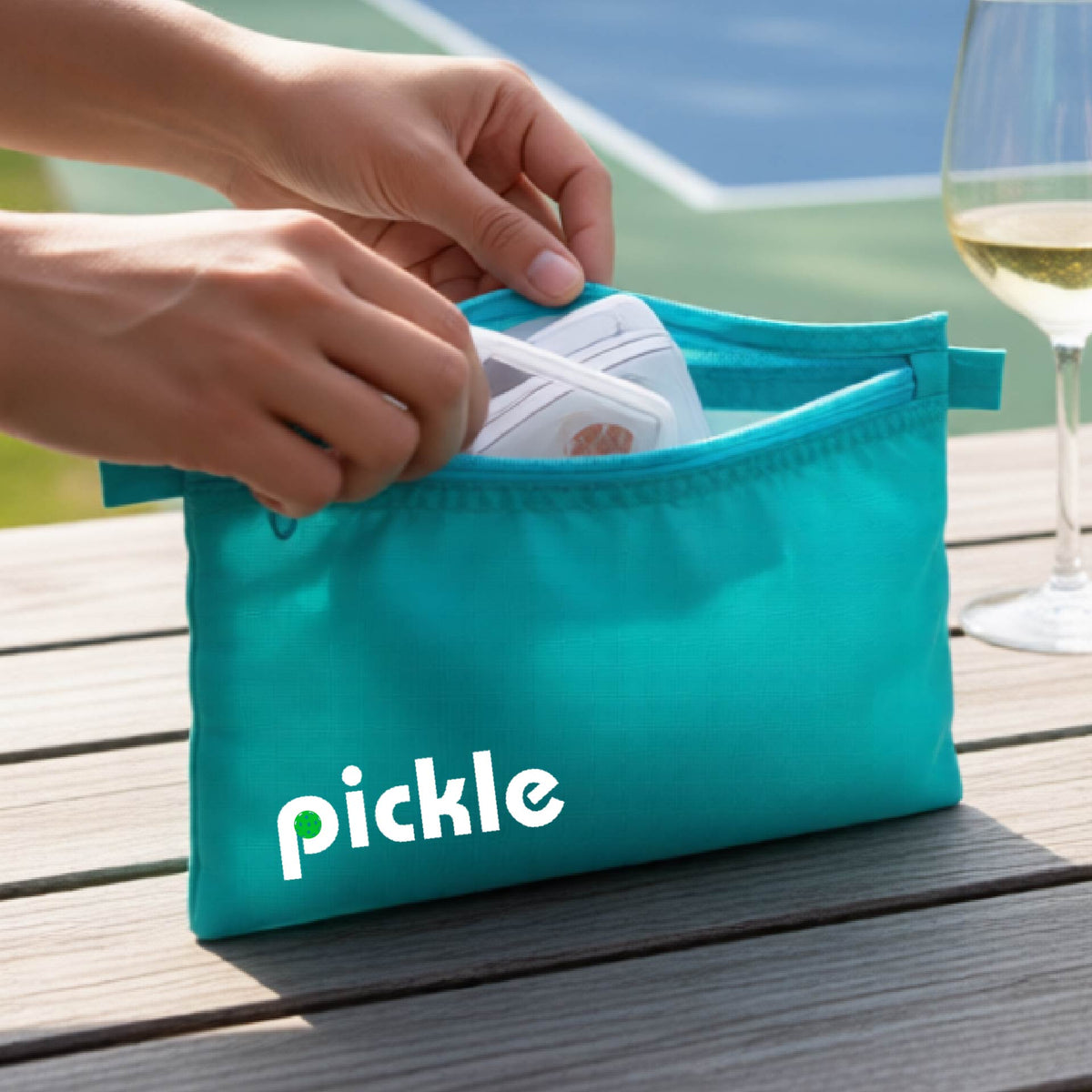 A close-up of a teal pickleball polyester cosmetic bag with a white graphic of ‘PICKLE’ with a green pickleball integrated into the letter P printed on the upper right corner, in front of a pickleball court. 