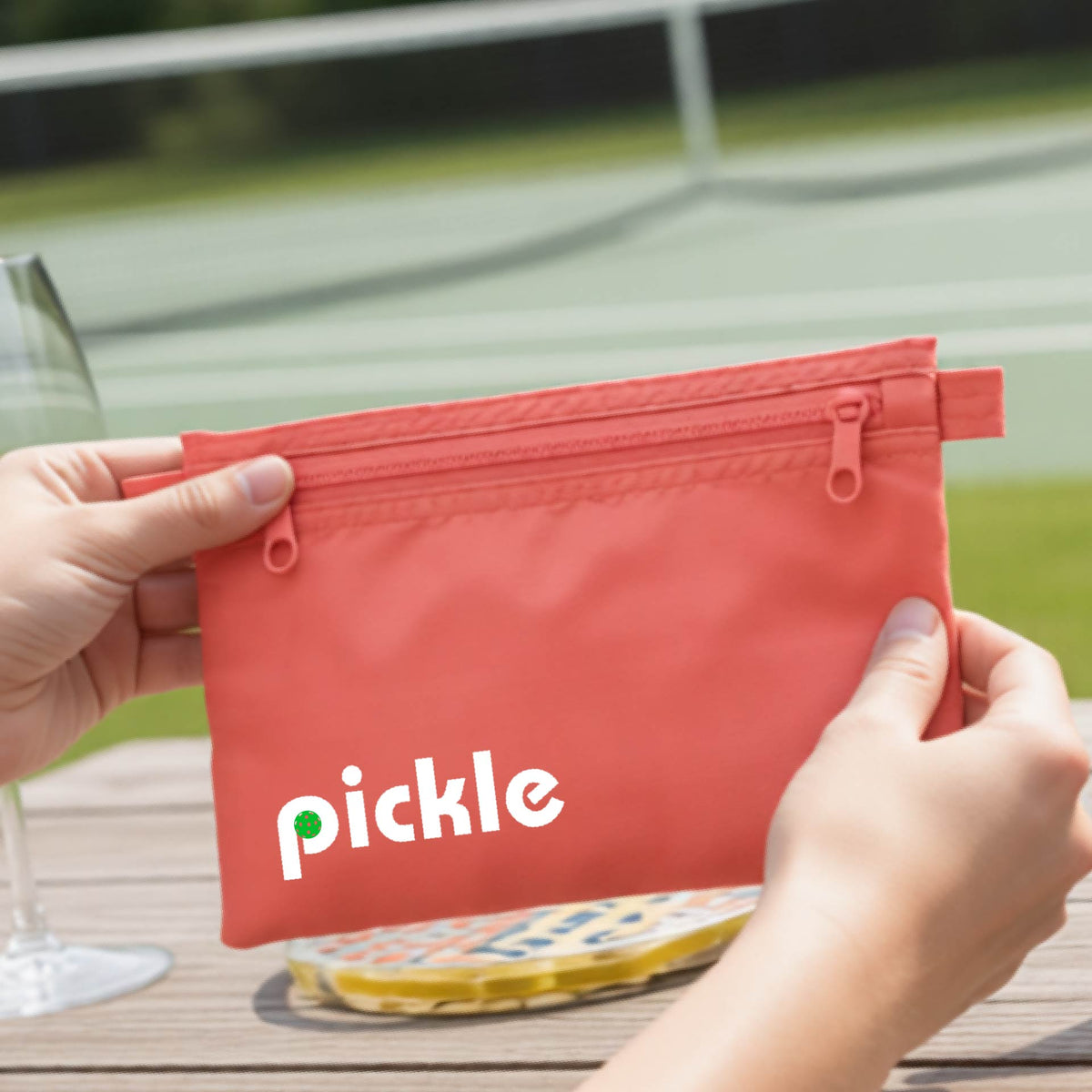 A close-up of a coral pickleball polyester cosmetic bag with a white graphic of ‘PICKLE’ with a green pickleball integrated into the letter P printed on the upper right corner, in front of a pickleball court. 