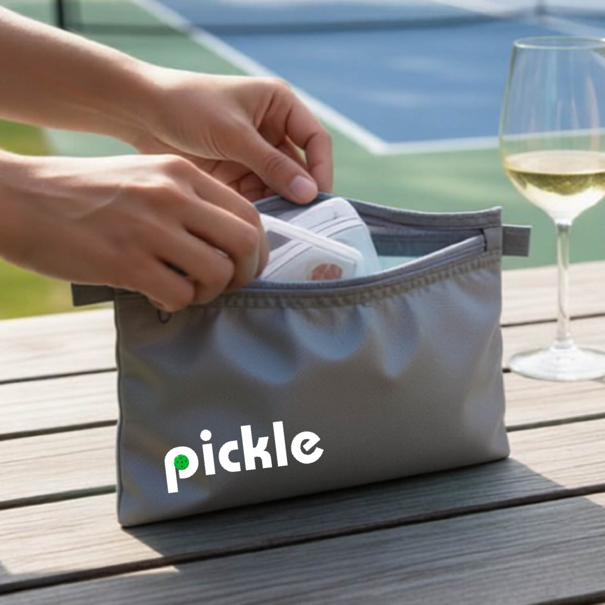 A close-up of a gray pickleball polyester cosmetic bag with a white graphic of ‘PICKLE’ with a green pickleball integrated into the letter P printed on the upper right corner, in front of a pickleball court. 