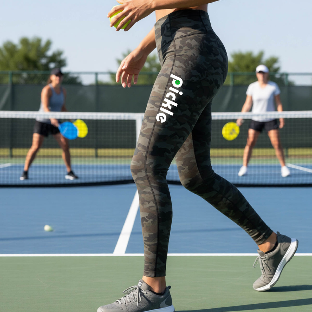 A close-up lifestyle shot of a woman on a pickleball court wearing high-waisted camo leggings with 'Pickle' printed in white down the side of her leg.