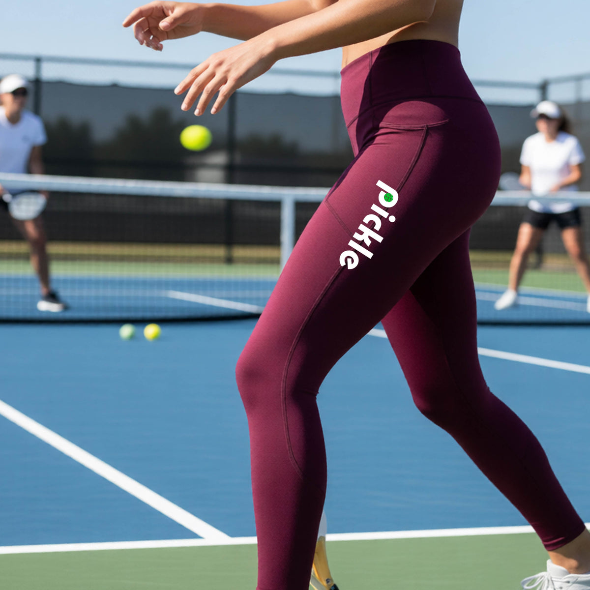 A close-up lifestyle shot of a woman on a pickleball court wearing high-waisted maroon leggings with 'Pickle' printed in white down the side of her leg.