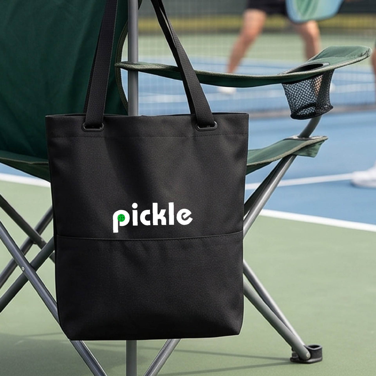 A black canvas tote bag with the word 'pickle' and a pickleball icon in the middle of the letter P printed in white across the front, on a chair in front of a pickleball court. 