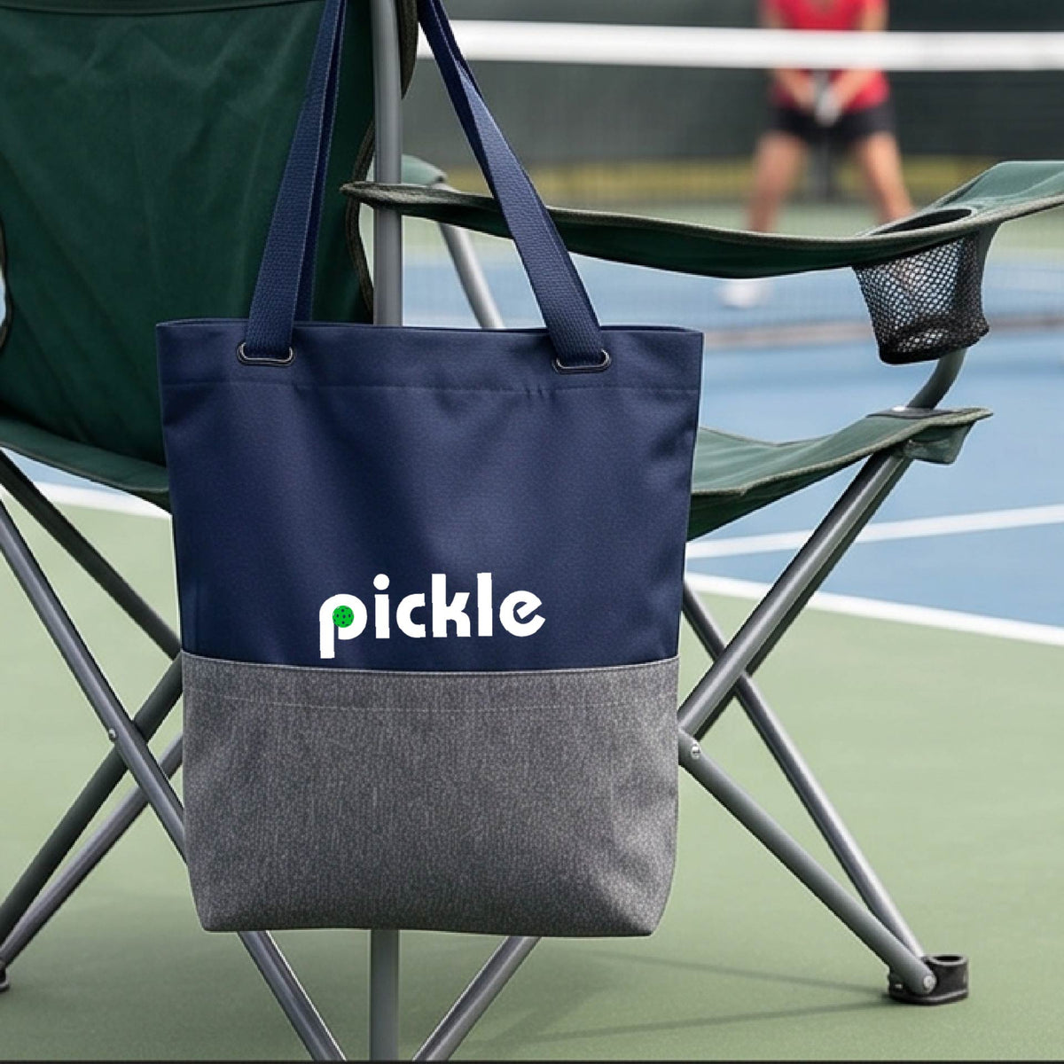 A blue and gray canvas tote bag with the word 'pickle' and a pickleball icon in the middle of the letter P printed in white across the front, on a chair in front of a pickleball court. 