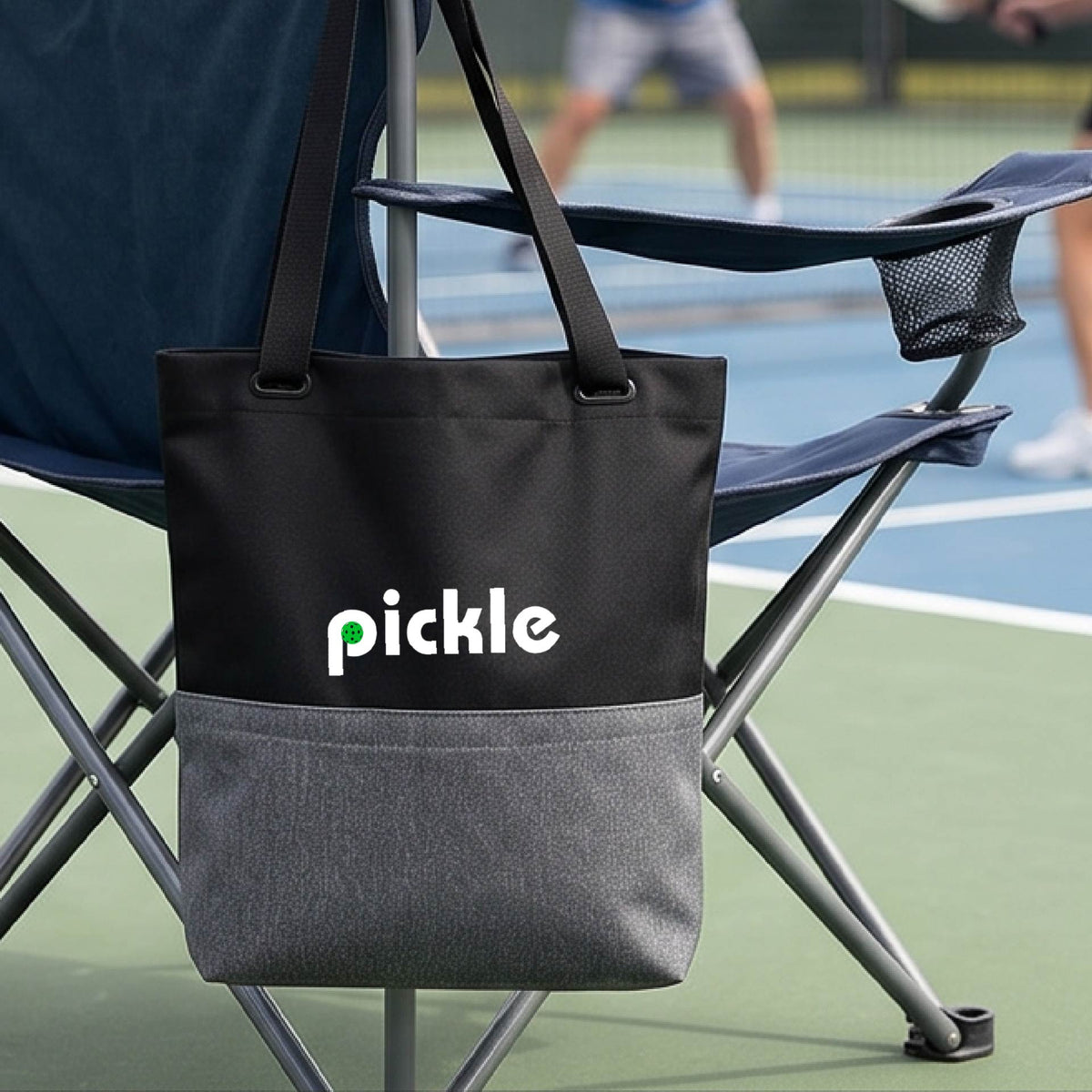 A gray black canvas tote bag with the word 'pickle' and a pickleball icon in the middle of the letter P printed in white across the front, on a chair in front of a pickleball court. 