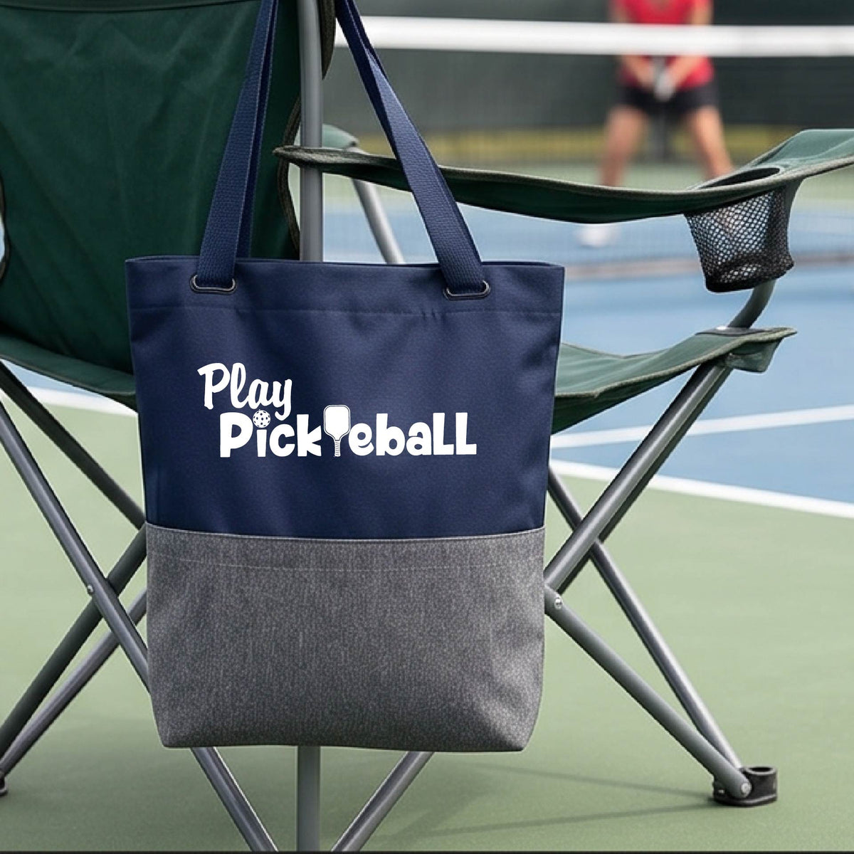 A navy blue and gray tote bag with white 'Play Pickleball' text hanging from a green folding chair on a pickleball court, with a blurred player in the background.