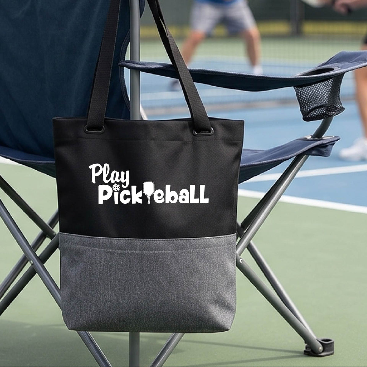 A black and gray tote bag with the text 'Play Pickleball' printed in white, hanging from a folding chair on a pickleball court with a blurred player in the background.