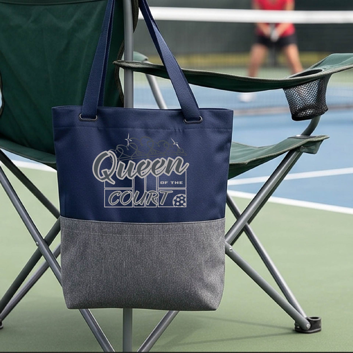 A navy blue canvas tote bag with a gray lower pocket, with 'Queen of the Court' pickleball motiff printed in white on the front, hanging from the armrest of a green folding chair on a pickleball court.