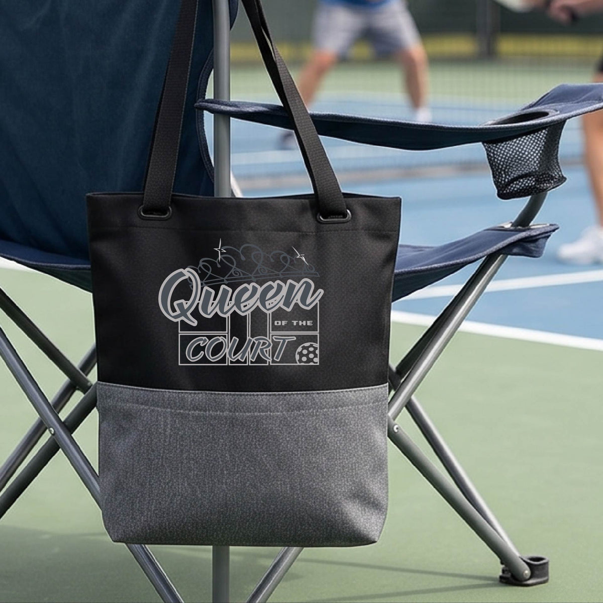 A black and gray tote bag with 'Queen of the Court' pickleball theme printed in white on the front, hanging from the armrest of a green folding chair on a pickleball court.