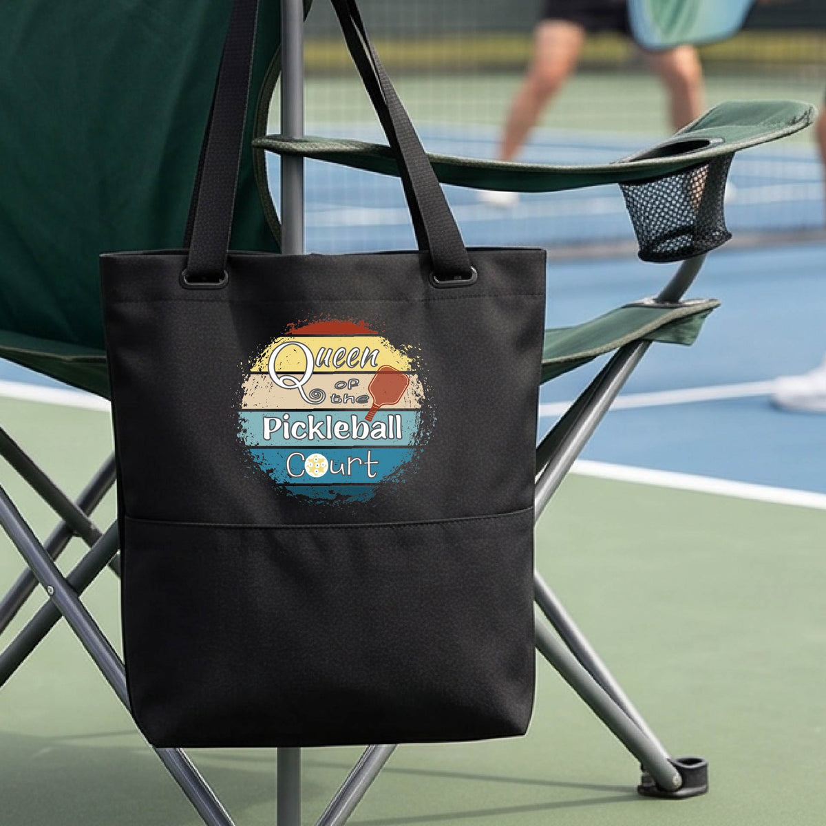A black tote bag  hangs from a folding chair on a pickleball court. The front pocket has a colorful circular graphic that reads 'Queen of the Pickleball Court' with a retro sunset design.