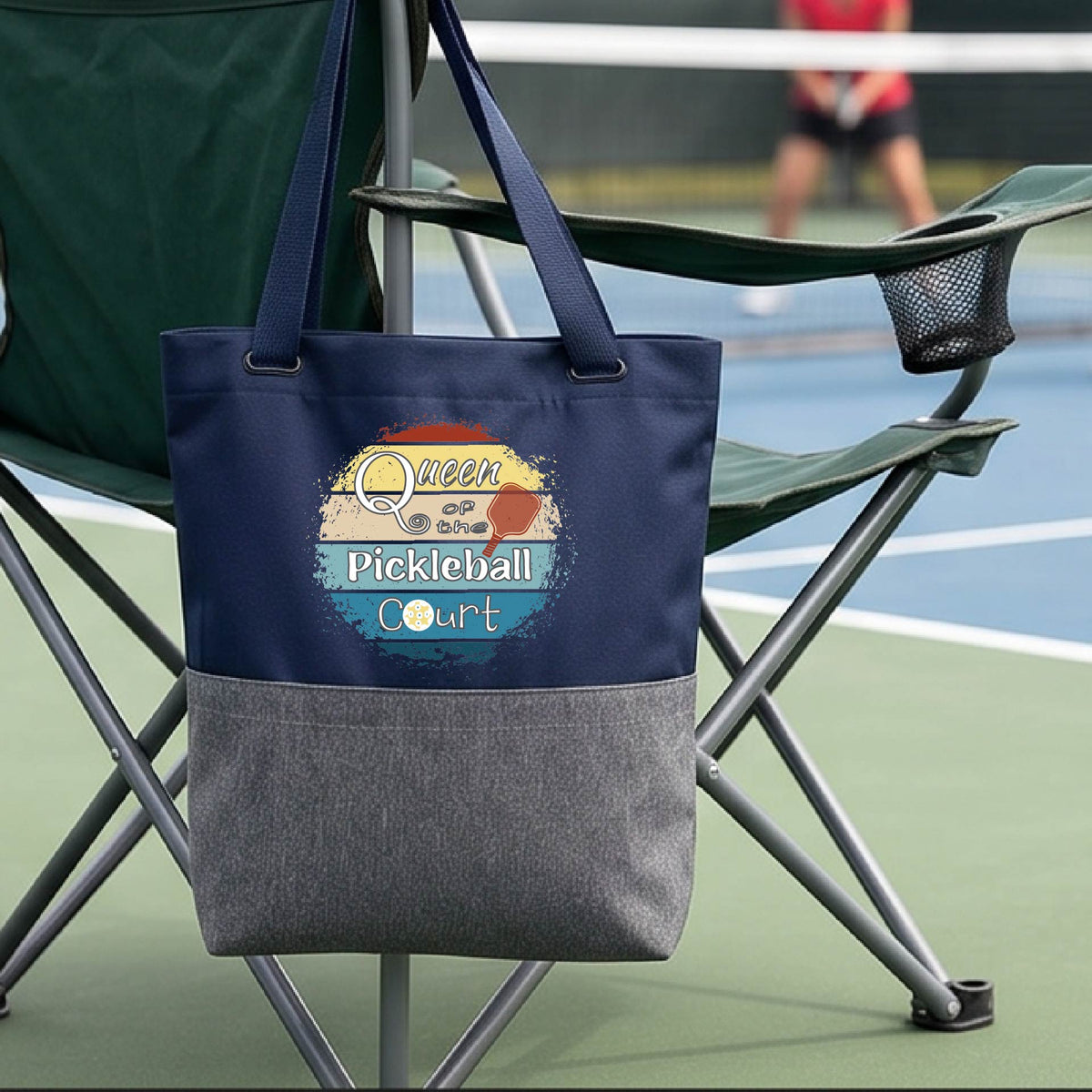 A navy blue canvas tote bag with a gray fabric bottom, hanging from the arm of a green folding chair on a pickleball court. The bag features a colorful retro-style graphic that reads 'Queen of the  Pickleball Court' with a retro paddle graphic.