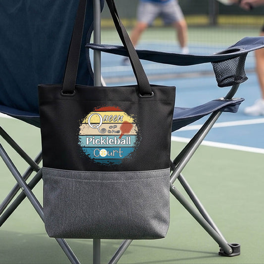 A black tote bag with a gray bottom hangs from a folding chair on a pickleball court. The front pocket has a colorful circular graphic that reads 'Queen of the Pickleball Court' with a retro sunset design.