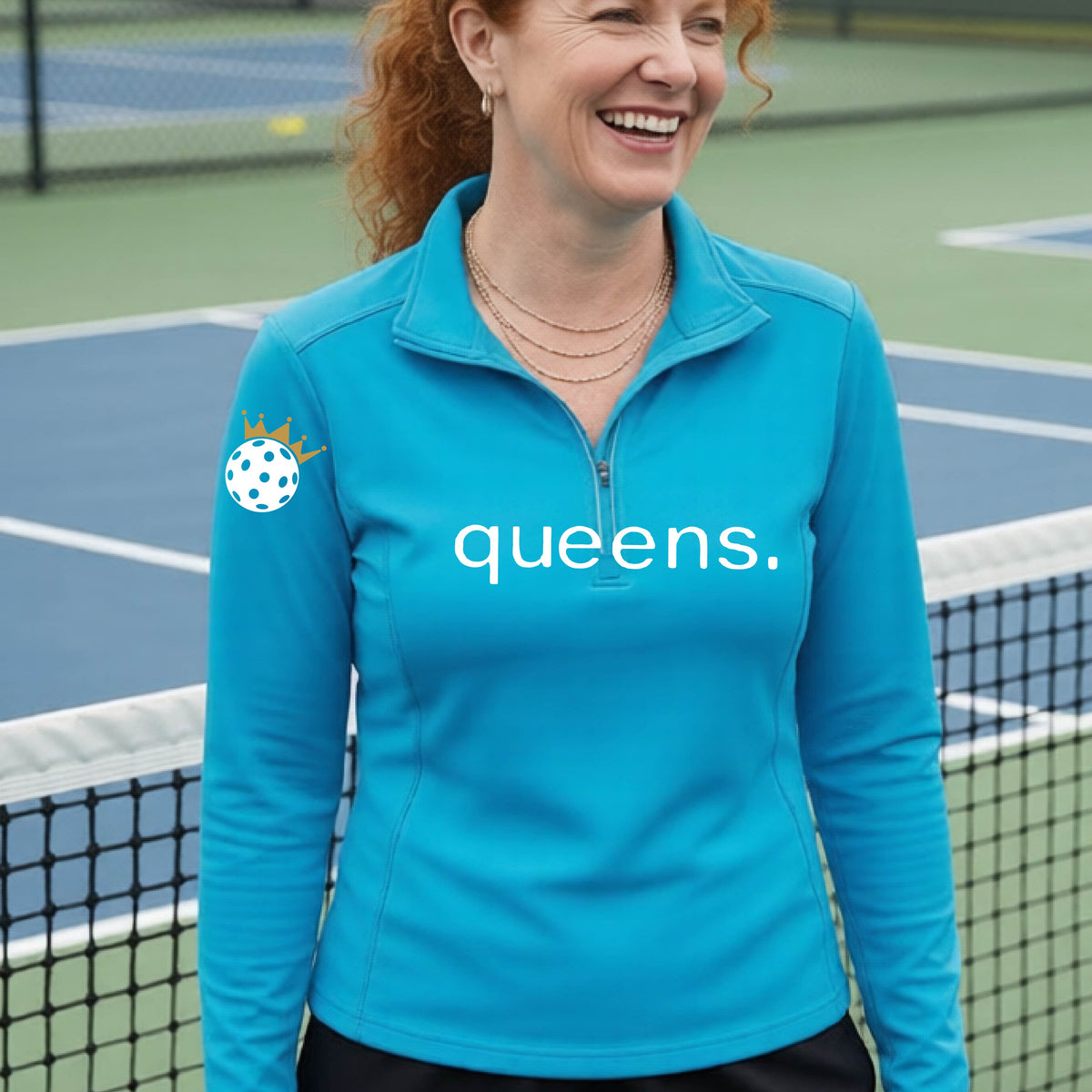 A woman smiling on a pickleball court wearing a bright blue long-sleeve quarter-zip top with 'queens.' printed in white across the chest and a white pickleball wearing a crown on the right sleeve.
