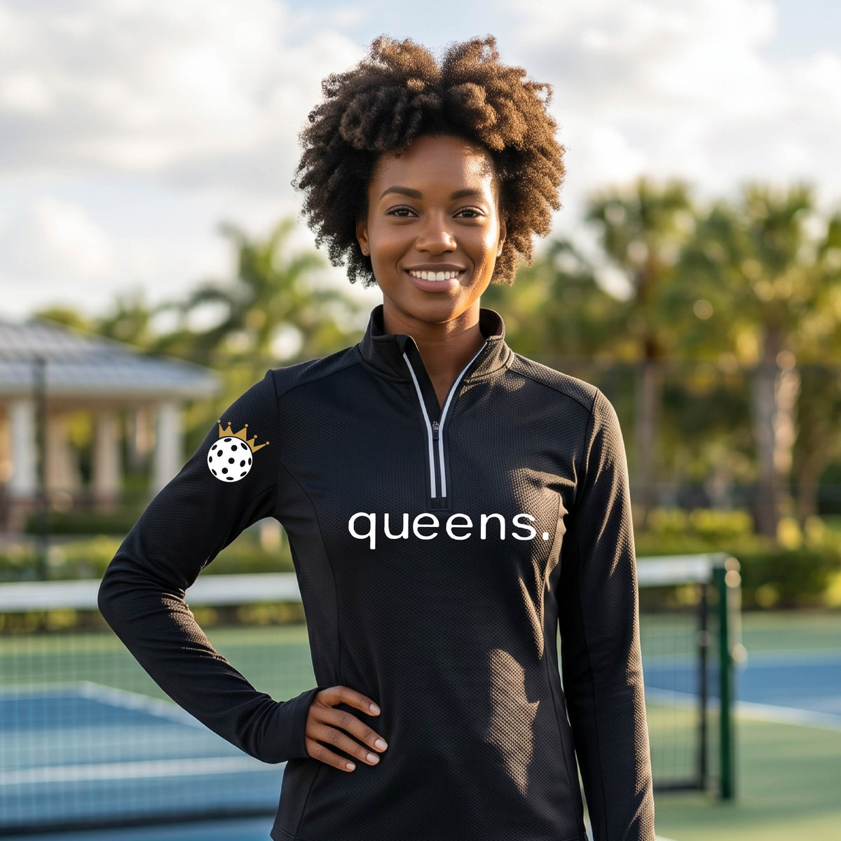 A woman stands on an outdoor pickkleball court wearing a black long-sleeve athletic quarter-zip top with a white 'queens.' wordmark across the chest and a small white pickleball wearing a gold crown logo on the sleeve.