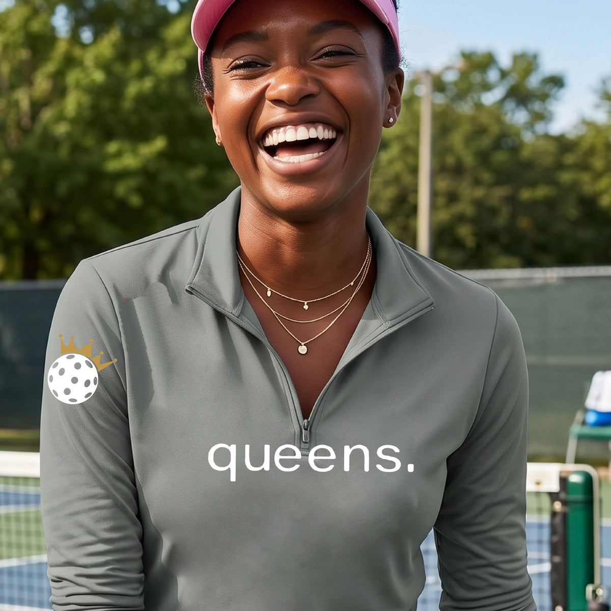 A woman smiling on a pickleball court wearing a gray long-sleeve quarter-zip top with 'queens.' printed in white across the chest and a white pickleball wearing a crown on the right sleeve.