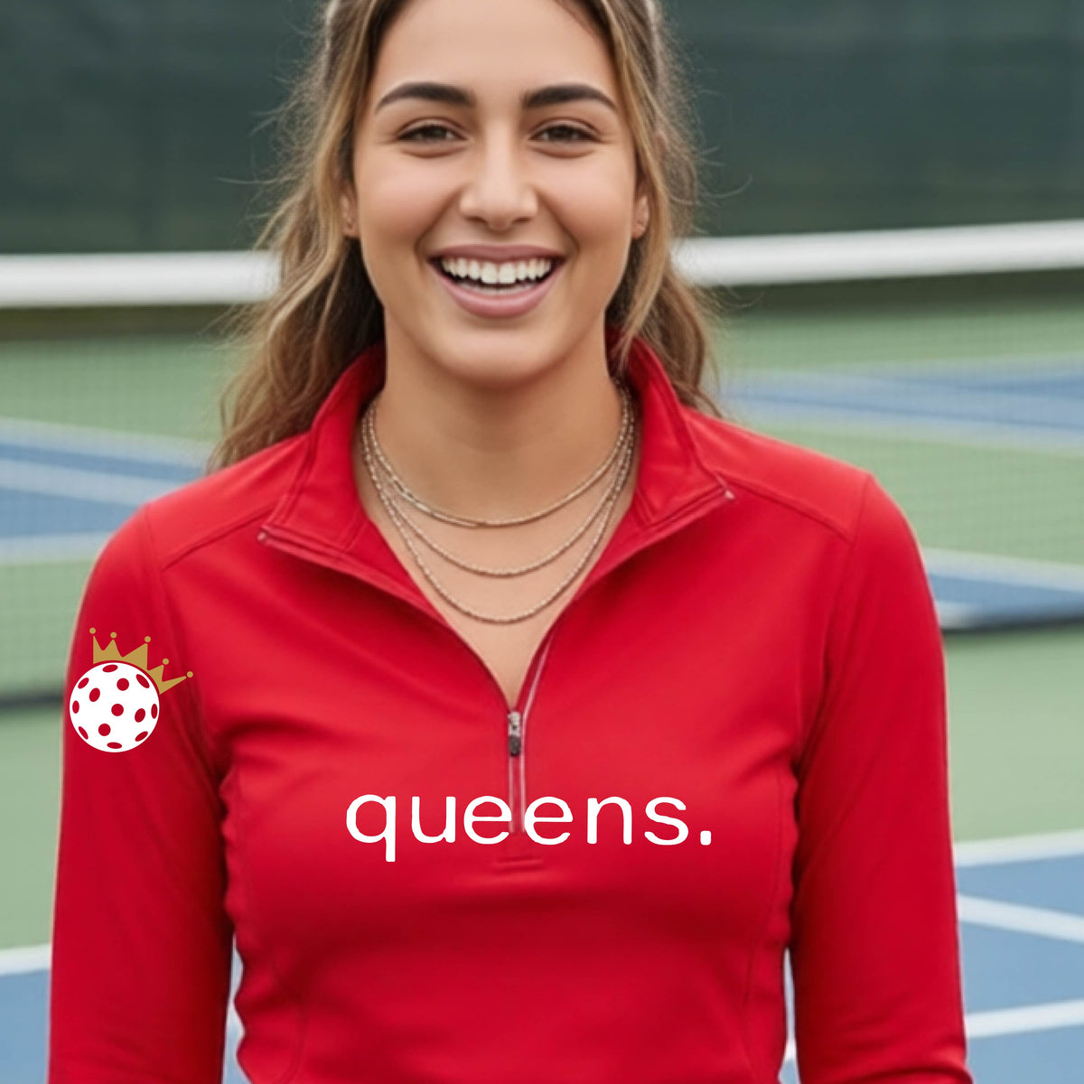 A woman smiling on a pickleball court wearing a red long-sleeve quarter-zip top with 'queens.' printed in white across the chest and a white pickleball wearing a crown on the right sleeve.