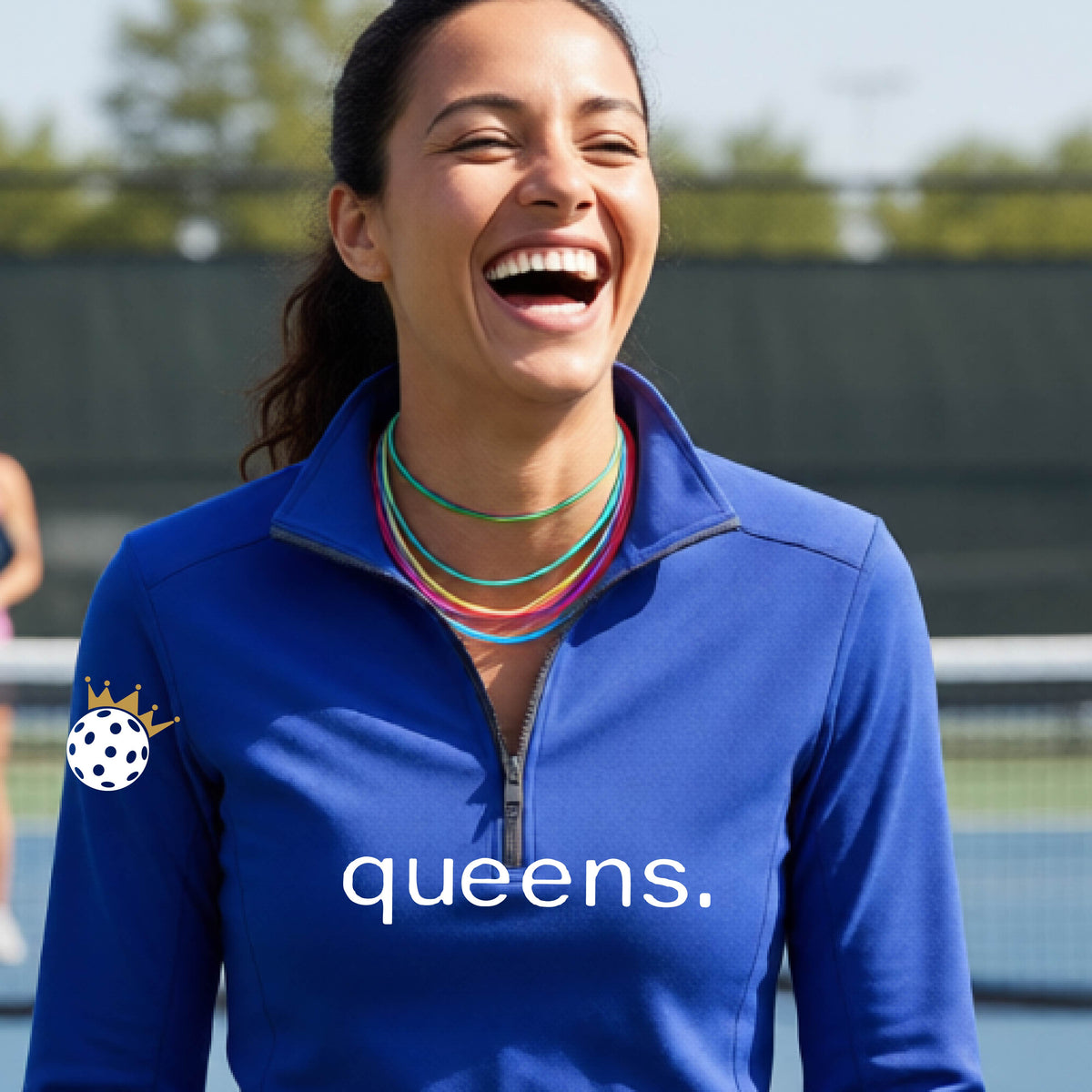 A woman smiling on a pickleball court wearing a roya blue long-sleeve quarter-zip top with 'queens.' printed in white across the chest and a white pickleball wearing a crown on the right sleeve.