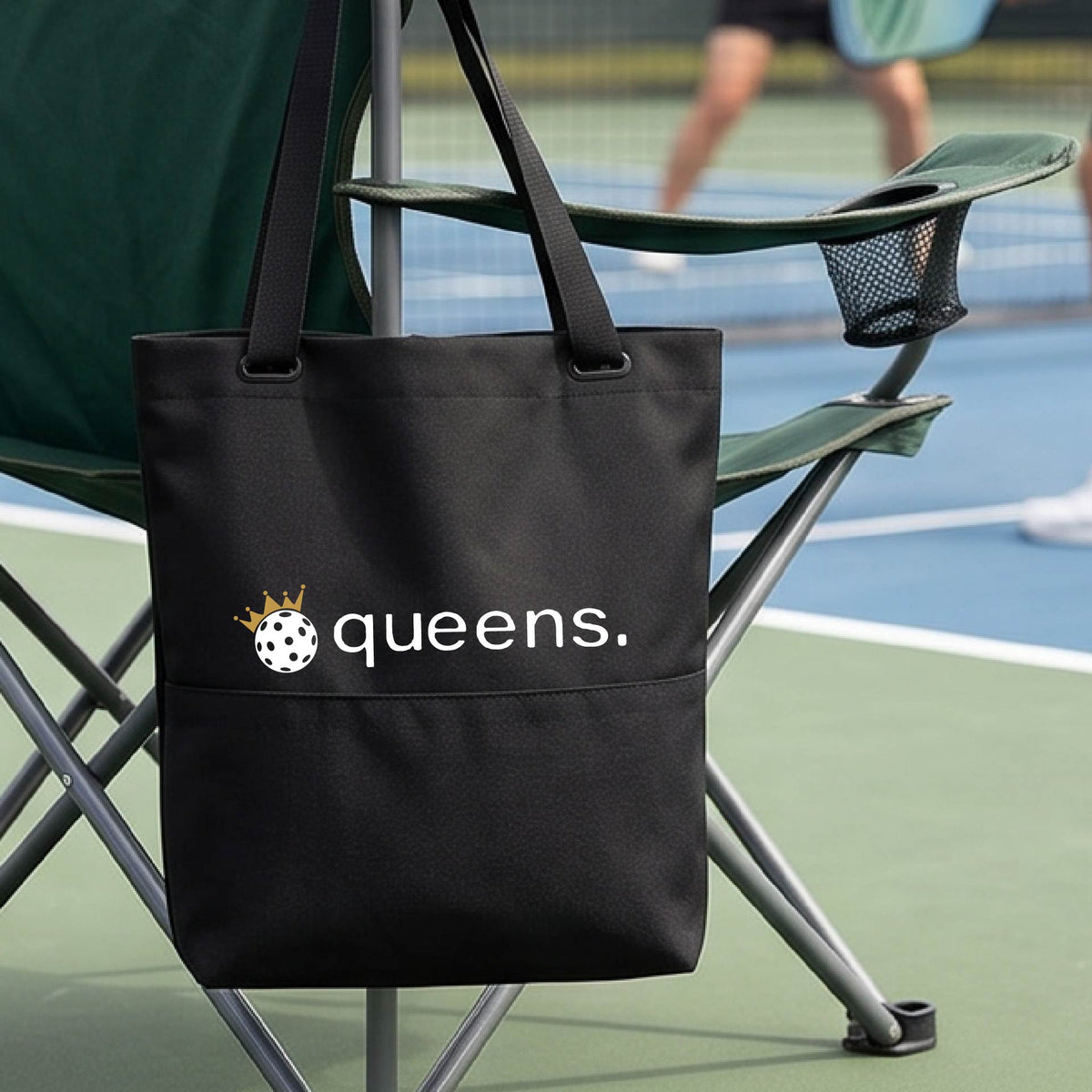 A black tote bag with the text 'queens.' and a gold crown on top of a pickleball logo, hanging from a folding chair on a pickleball court.