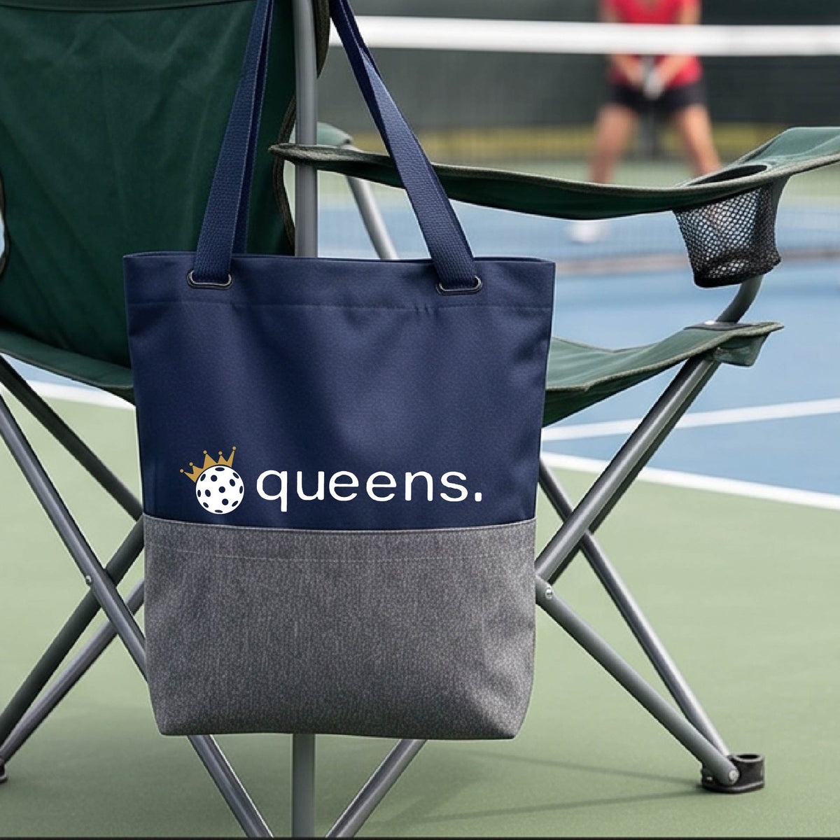 A blue and gray tote bag with the text 'queens.' and a gold crown on top of a pickleball logo, hanging from a folding chair on a pickleball court.