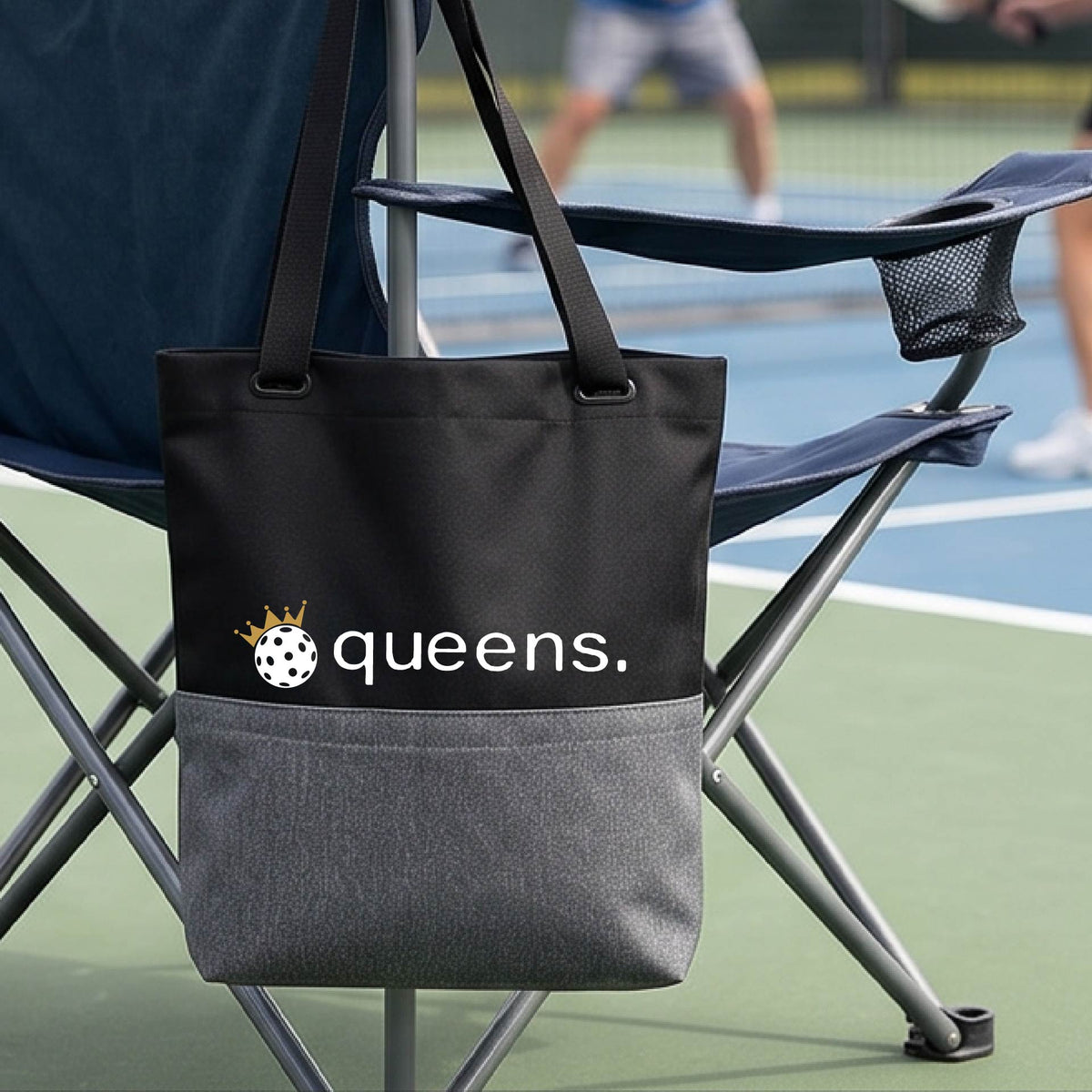 A black and gray tote bag with the text 'queens.' and a gold crown on top of a pickleball logo, hanging from a folding chair on a pickleball court.