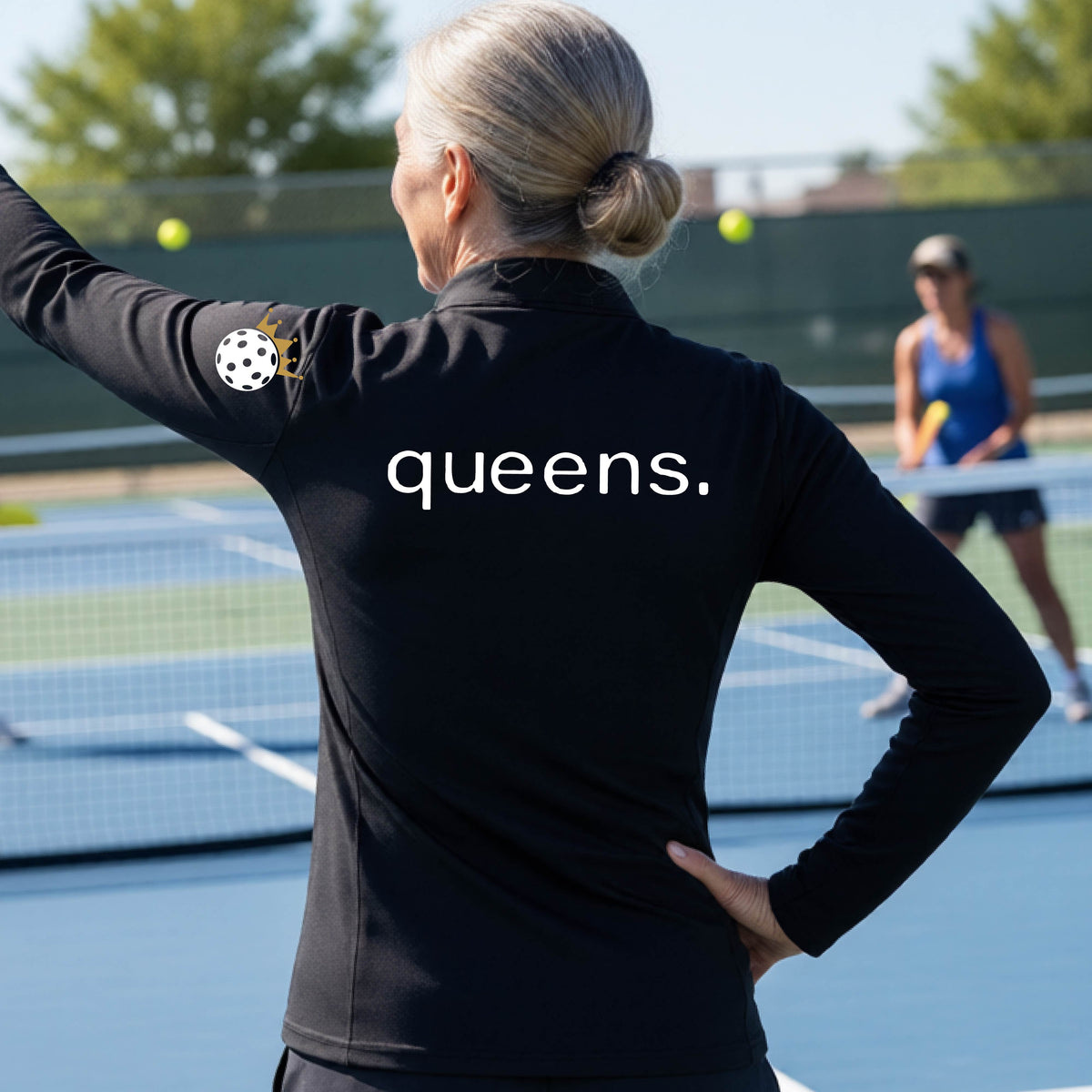 A woman on a pickleball court seen from behind, wearing a black long-sleeve athletic top with the word 'queens.' printed across the back and a white pickleball wearing a gold crown on her left sleeve. 