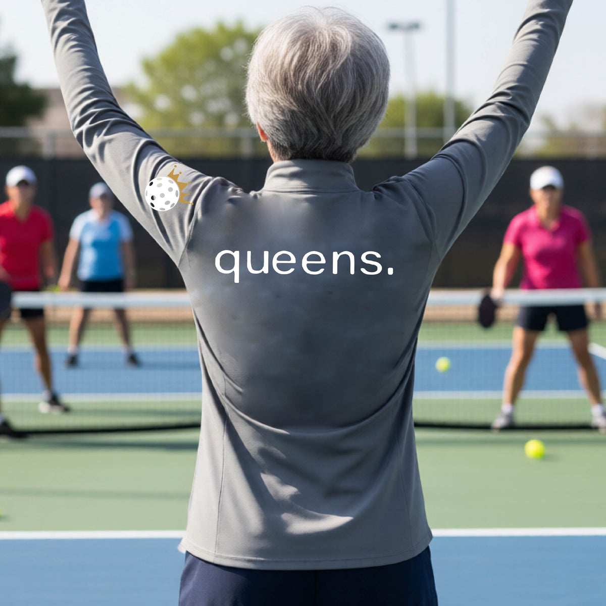 A woman on a pickleball court seen from behind, wearing a gray long-sleeve athletic top with the word 'queens.' printed across the back and a white pickleball wearing a gold crown on her left sleeve. 