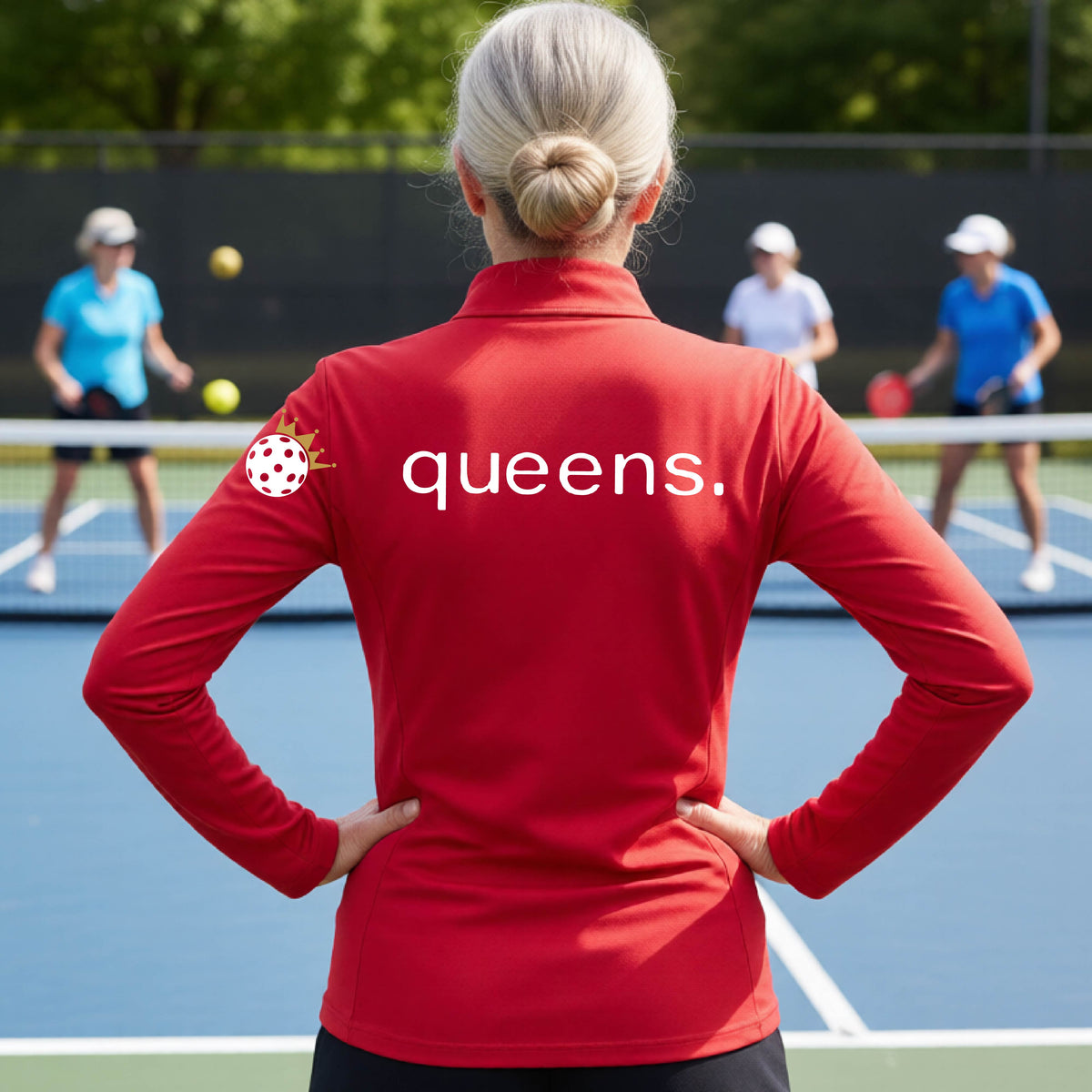 A woman on a pickleball court seen from behind, wearing a red long-sleeve athletic top with the word 'queens.' printed across the back and a white pickleball wearing a gold crown on her left sleeve. 