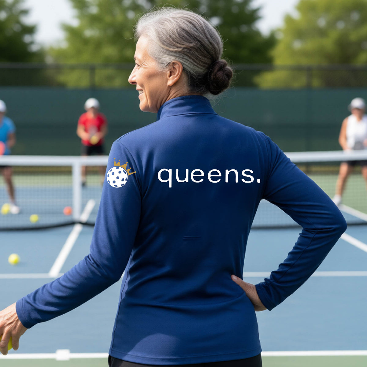 A woman on a pickleball court seen from behind, wearing a royal blue long-sleeve athletic top with the word 'queens.' printed across the back and a white pickleball wearing a gold crown on her left sleeve. 