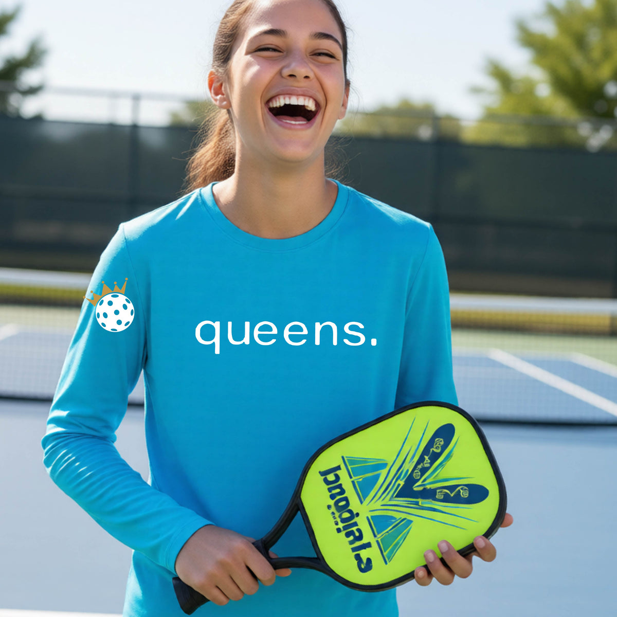 A woman laughing on a pickleball court wearing a bright blue long-sleeve shirt with 'queens.' printed in white across the chest and a small pickleball with a gold crown on the left sleeve.