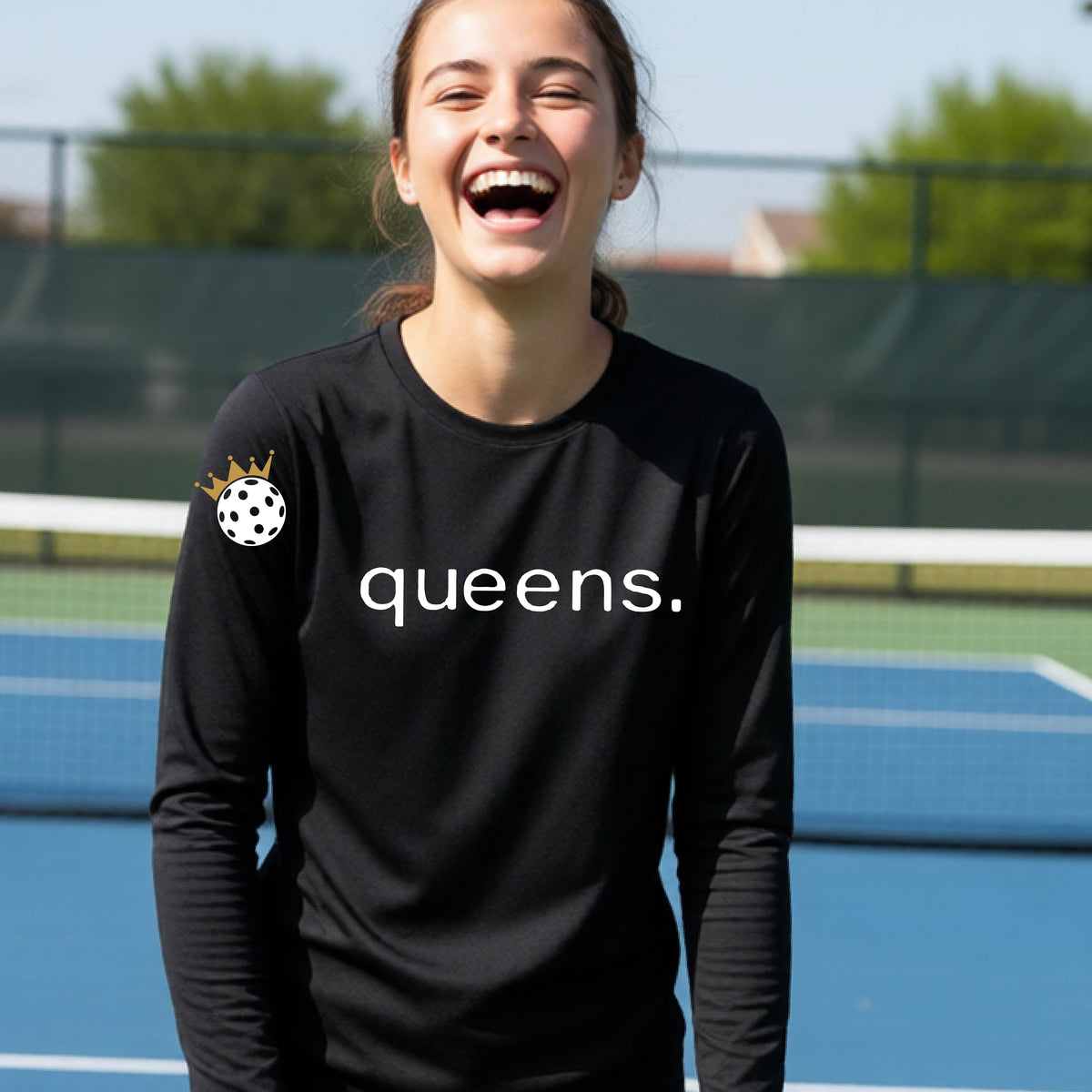 A woman laughing on a pickleball court wearing a black long-sleeve shirt with 'queens.' printed in white across the chest and a small pickleball with a gold crown on the left sleeve.