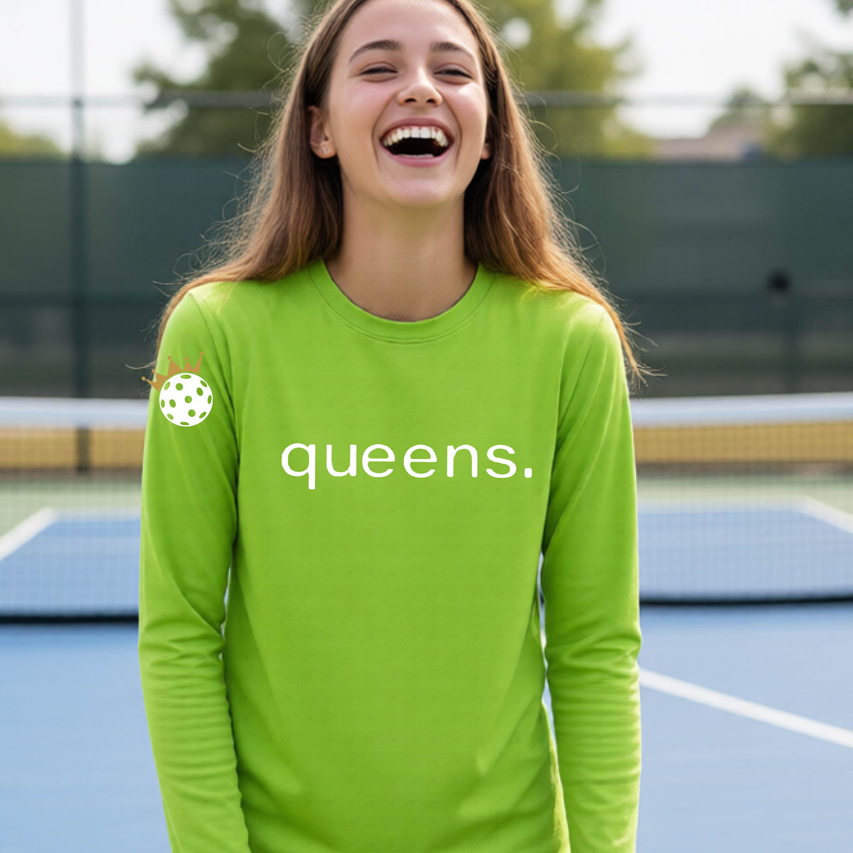 A woman laughing on a pickleball court wearing a lime green long-sleeve shirt with 'queens.' printed in white across the chest and a small pickleball with a gold crown on the left sleeve.