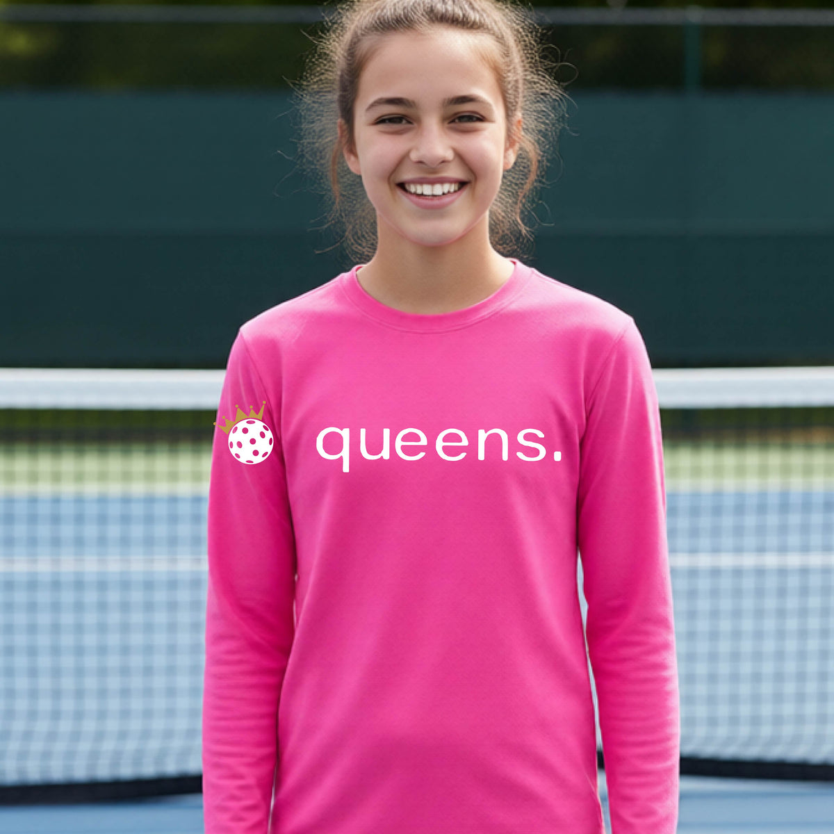 A woman laughing on a pickleball court wearing a pink long-sleeve shirt with 'queens.' printed in white across the chest and a small pickleball with a gold crown on the left sleeve.