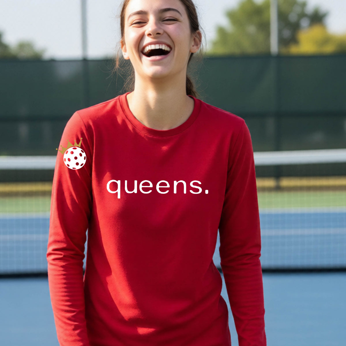 A woman laughing on a pickleball court wearing a red long-sleeve shirt with 'queens.' printed in white across the chest and a small pickleball with a gold crown on the left sleeve.