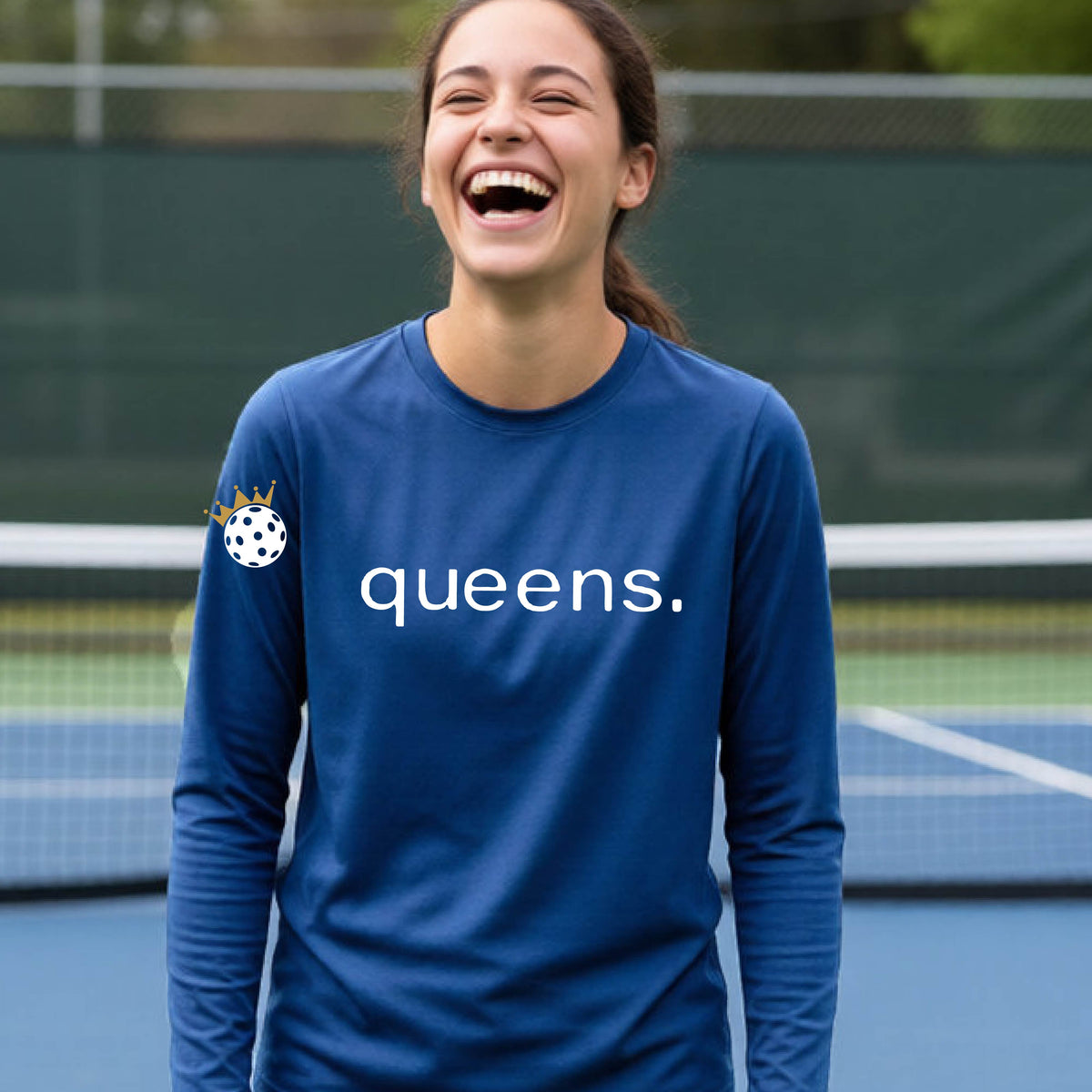 A woman laughing on a pickleball court wearing a royal blue long-sleeve shirt with 'queens.' printed in white across the chest and a small pickleball with a gold crown on the left sleeve.
