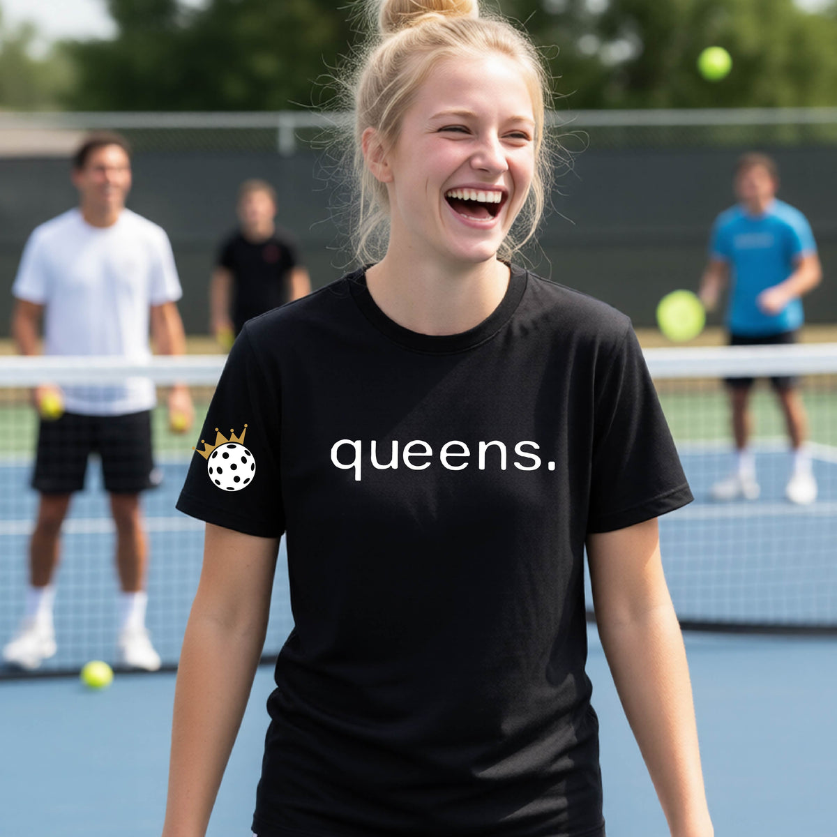A young woman laughing on a picleball court, wearing a black t-shirt with the word 'queens.' printed in white across the chest and a small white pickleball wearing a gold crown on the left sleeve.
