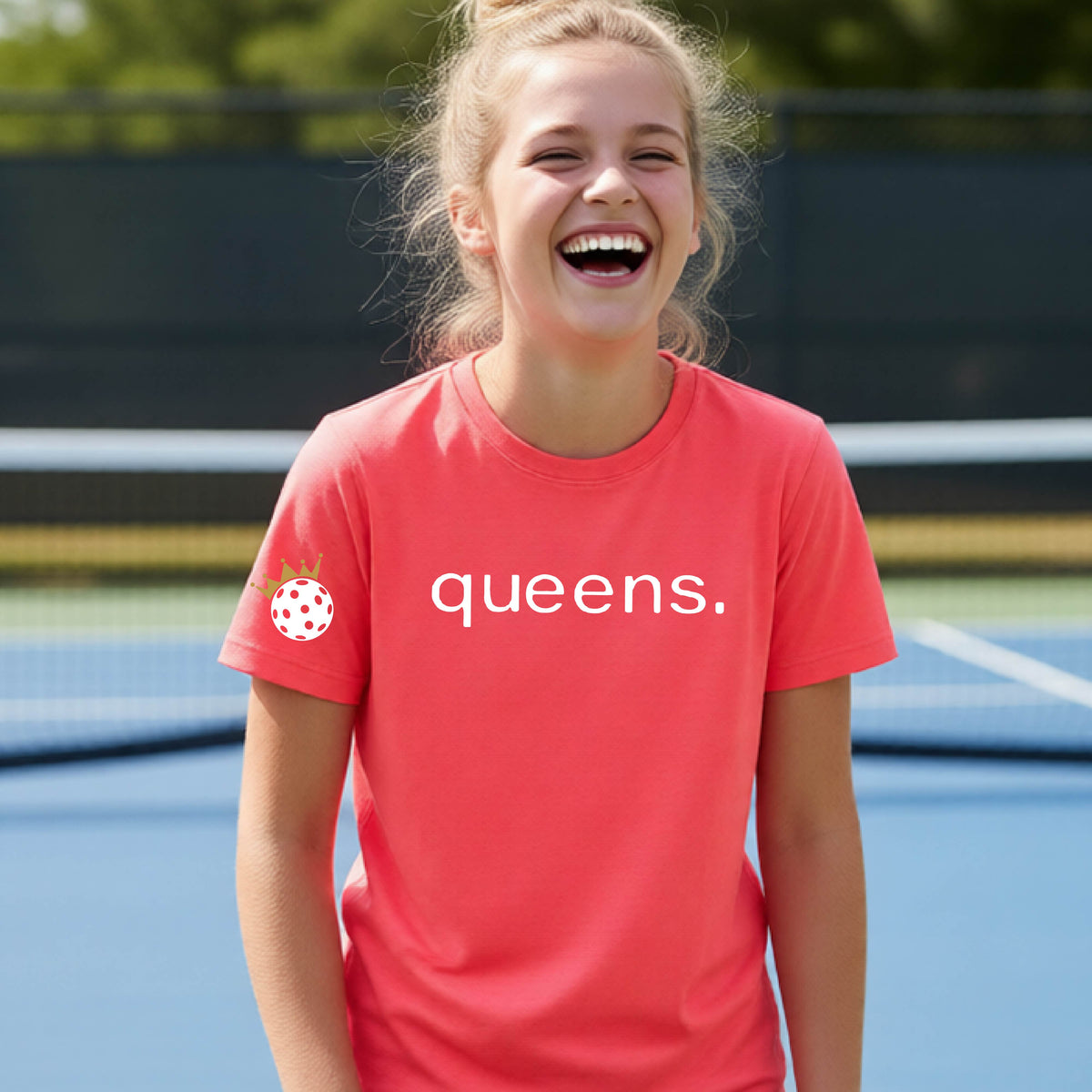 A young woman laughing on a picleball court, wearing a coral pink t-shirt with the word 'queens.' printed in white across the chest and a small white pickleball wearing a gold crown on the left sleeve.