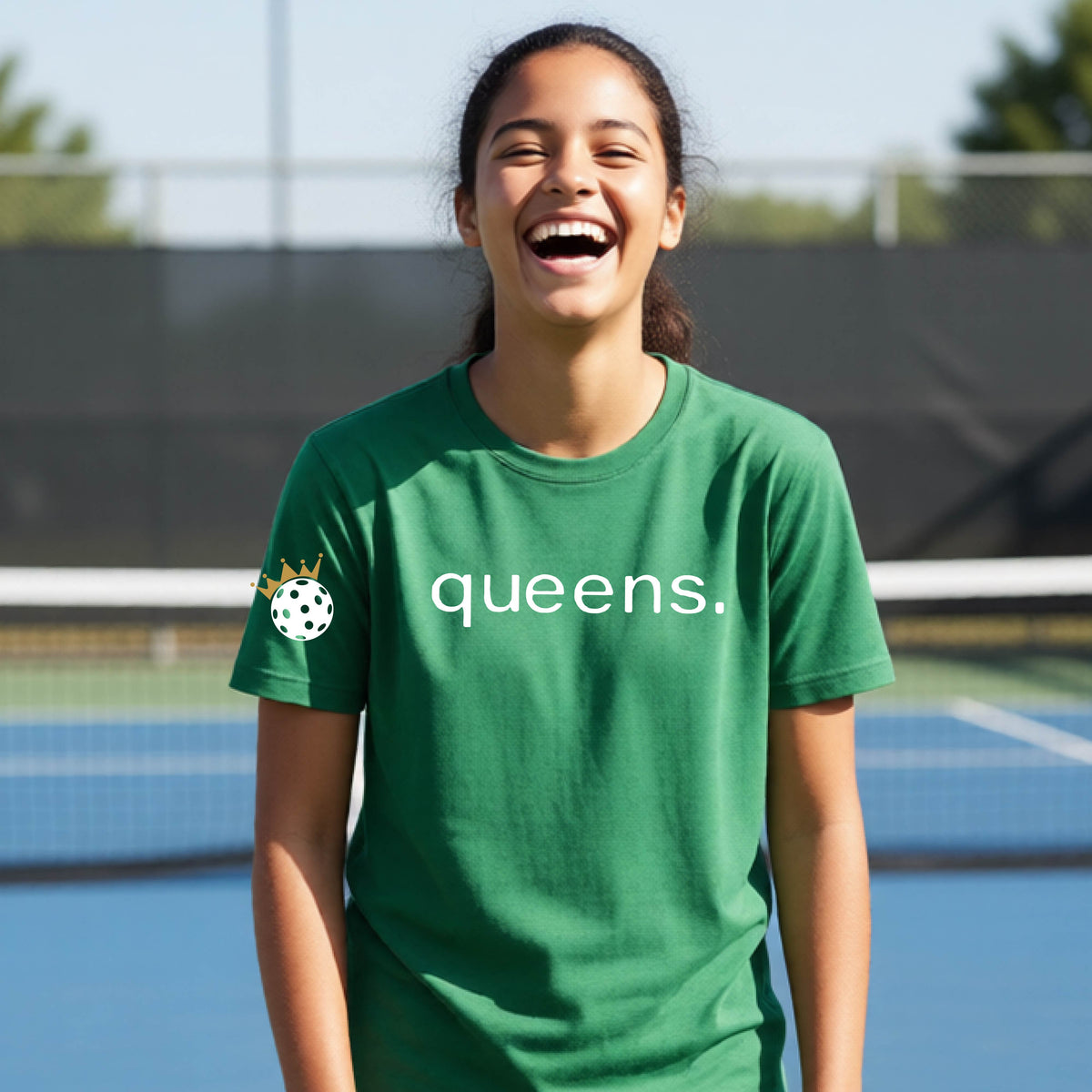 A young woman laughing on a picleball court, wearing a green t-shirt with the word 'queens.' printed in white across the chest and a small white pickleball wearing a gold crown on the left sleeve.
