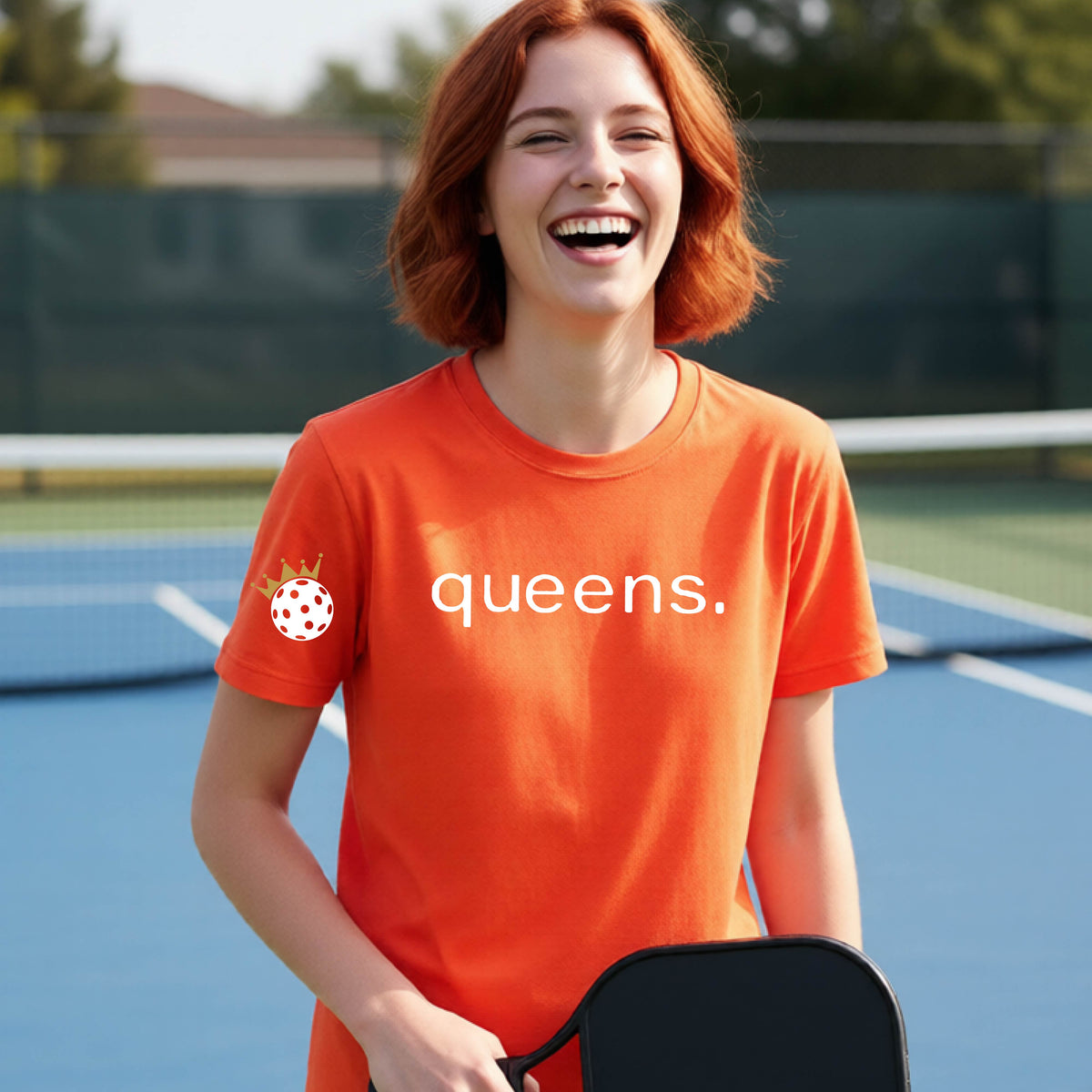 A young woman laughing on a picleball court, wearing an orange t-shirt with the word 'queens.' printed in white across the chest and a small white pickleball wearing a gold crown on the left sleeve.