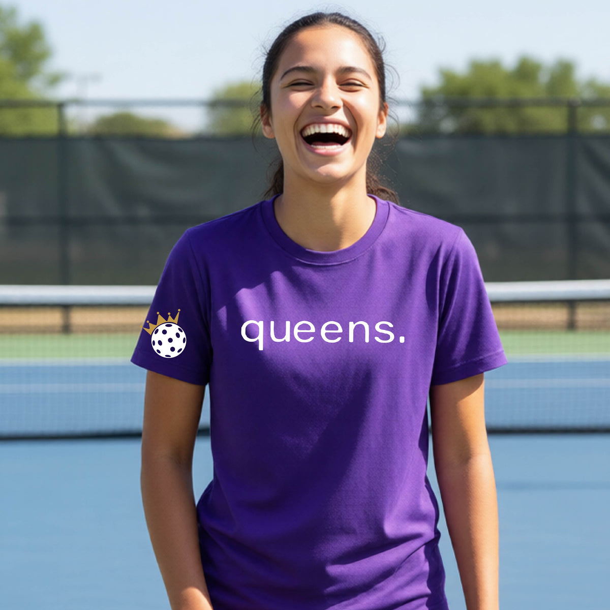 A young woman laughing on a picleball court, wearing a purple t-shirt with the word 'queens.' printed in white across the chest and a small white pickleball wearing a gold crown on the left sleeve.