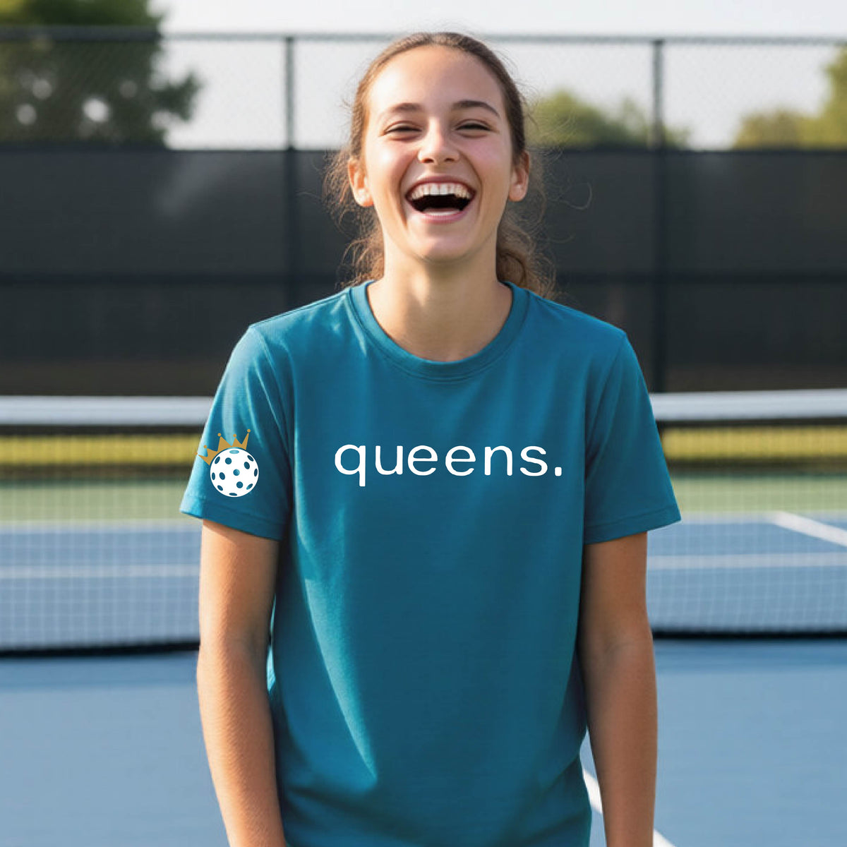 A young woman laughing on a picleball court, wearing a teal t-shirt with the word 'queens.' printed in white across the chest and a small white pickleball wearing a gold crown on the left sleeve.