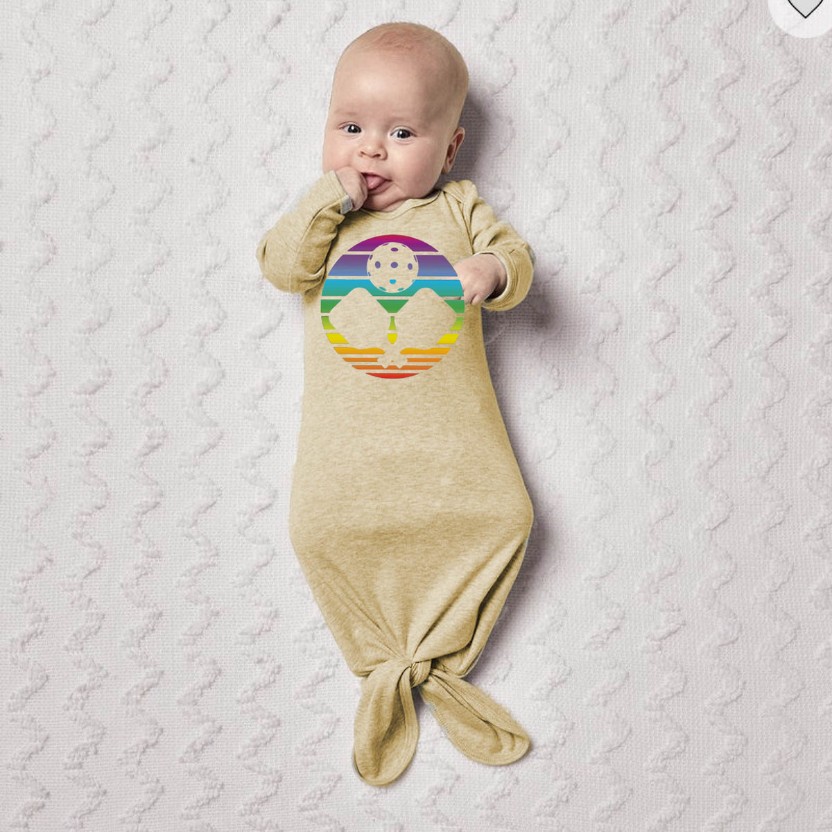 A baby lying on a white blanket wearing a beige sleep sack with a colorful rainbow pickleball paddle graphic.