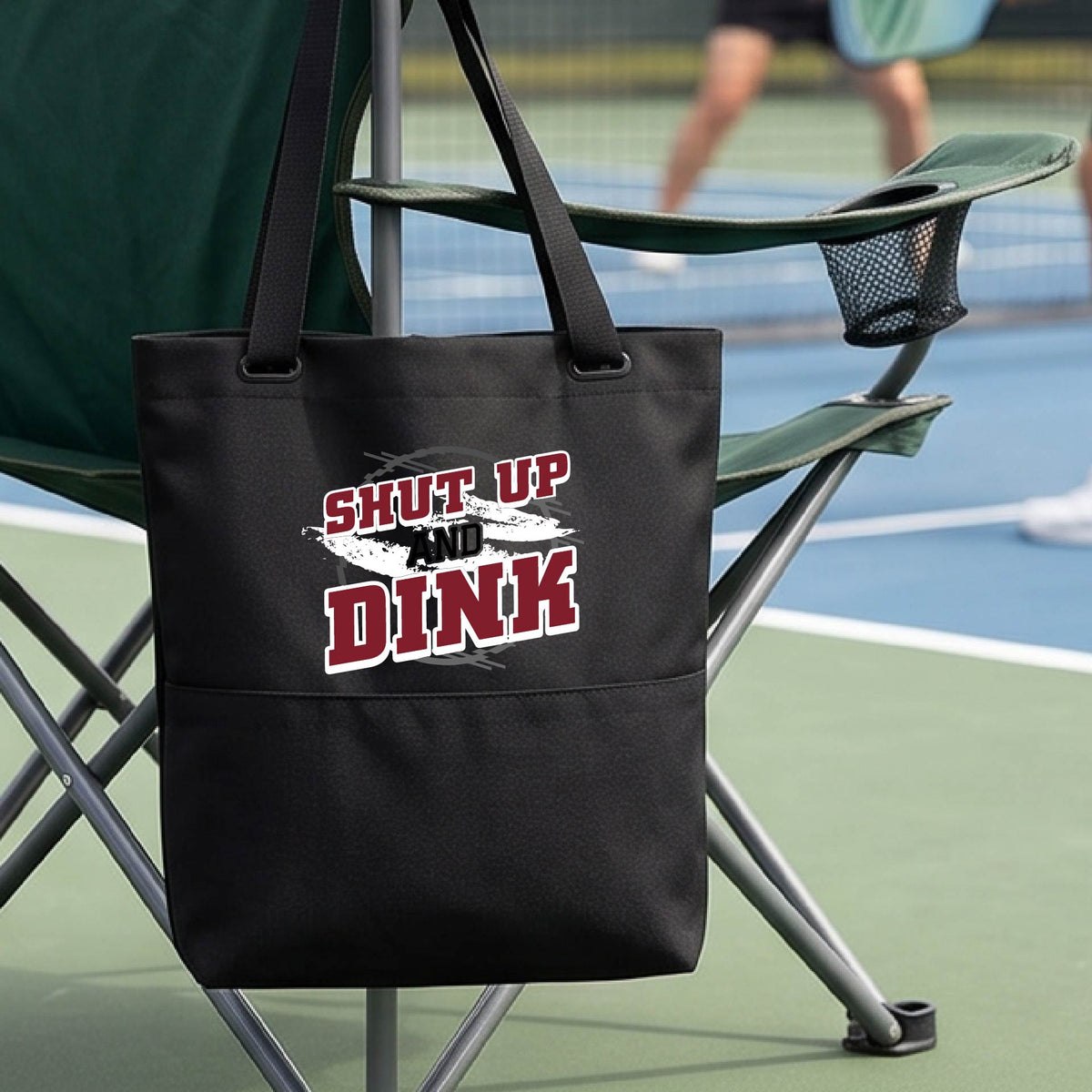 A close up of a black medium size tote bag with a white, gray and black graphic of the text: ‘Shut Up and Dink, on a pickleball court.