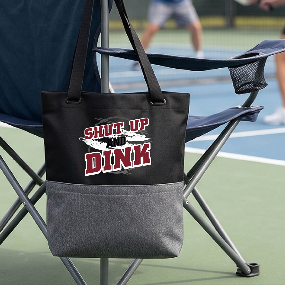 A close up of a black and gray medium size tote bag with a white, gray and black graphic of the text: ‘Shut Up and Dink, on a pickleball court.