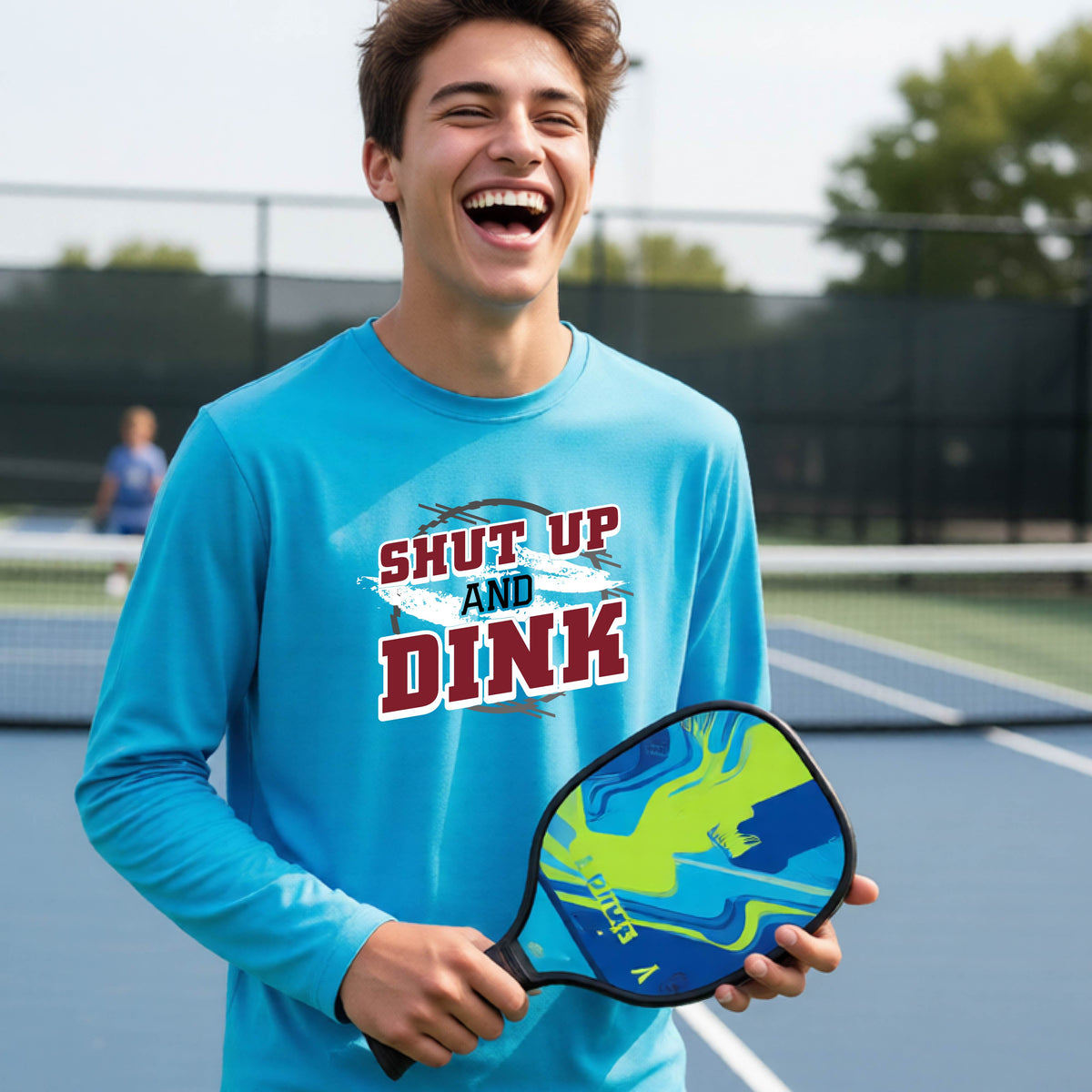 A young person on a pickleball court wearing a blue long-sleeve t-shirt with a white, gray and black graphic of the text:  ‘Shut Up and Dink, standing on a pickleball court.