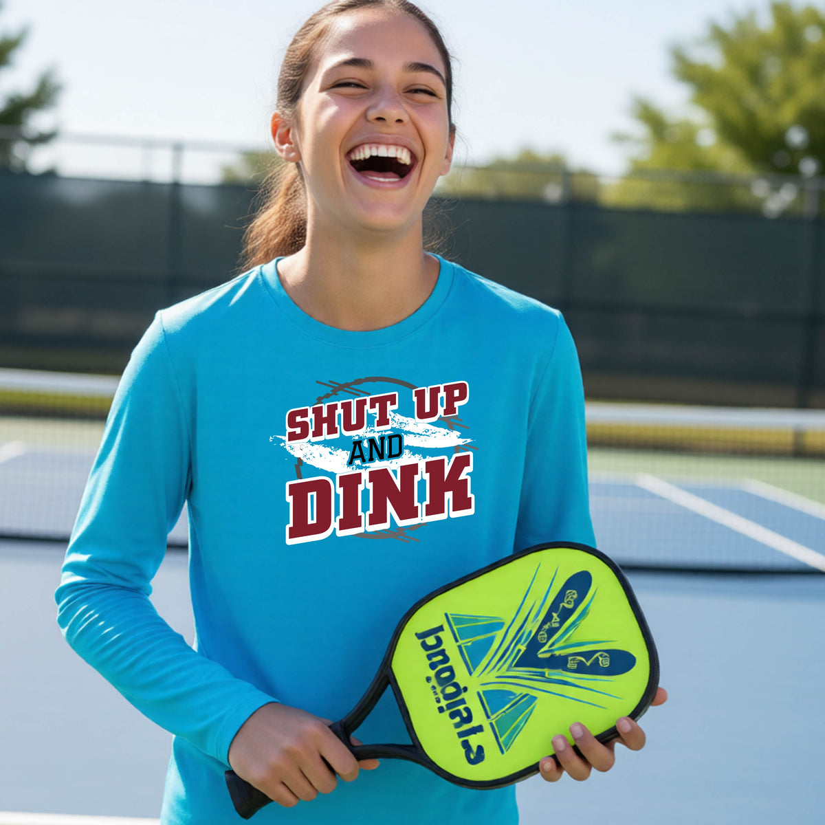 A young person on a pickleball court wearing a bright blue long-sleeve t-shirt with a white, gray and black graphic of the text:  ‘Shut Up and Dink, standing on a pickleball court.