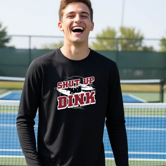A young person on a pickleball court wearing a black long-sleeve t-shirt with a white, gray and black graphic of the text:  ‘Shut Up and Dink, standing on a pickleball court.