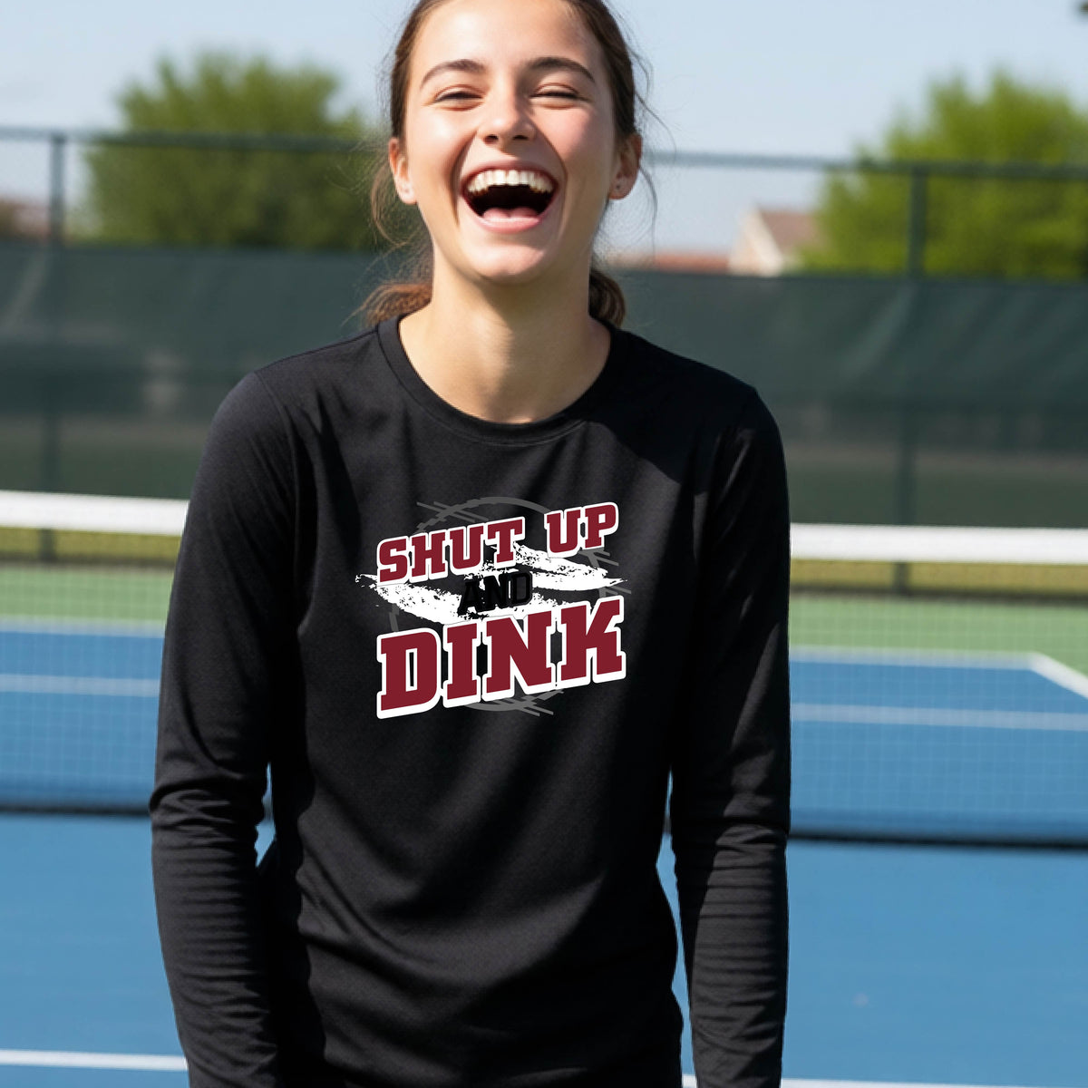 A young person on a pickleball court wearing a black long-sleeve t-shirt with a white, gray and black graphic of the text:  ‘Shut Up and Dink, standing on a pickleball court.