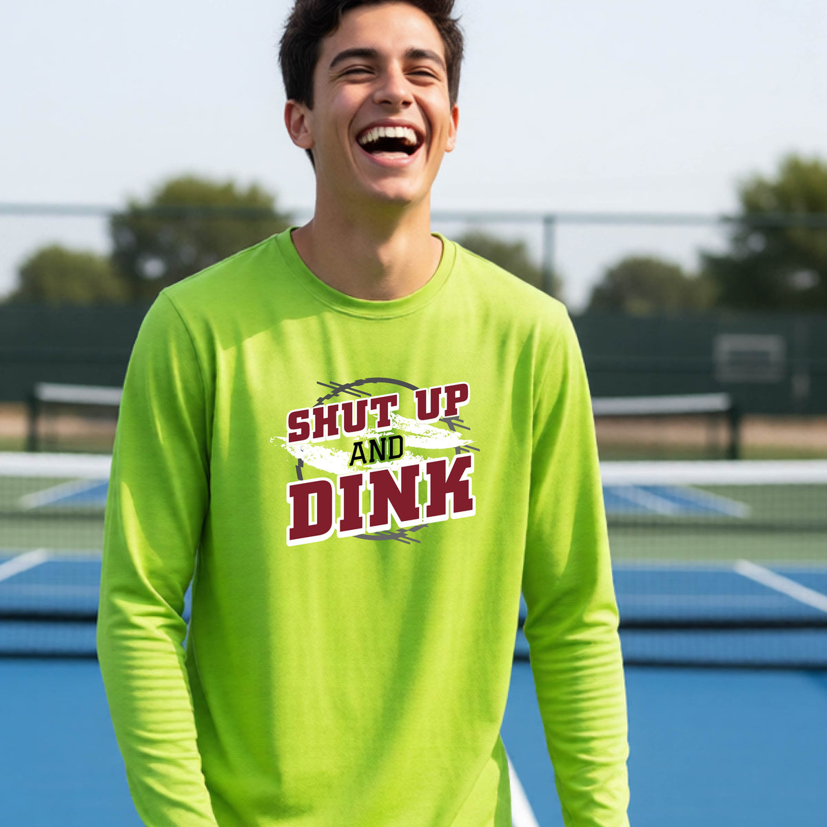 A young person on a pickleball court wearing a lime green long-sleeve t-shirt with a white, gray and black graphic of the text:  ‘Shut Up and Dink, standing on a pickleball court.