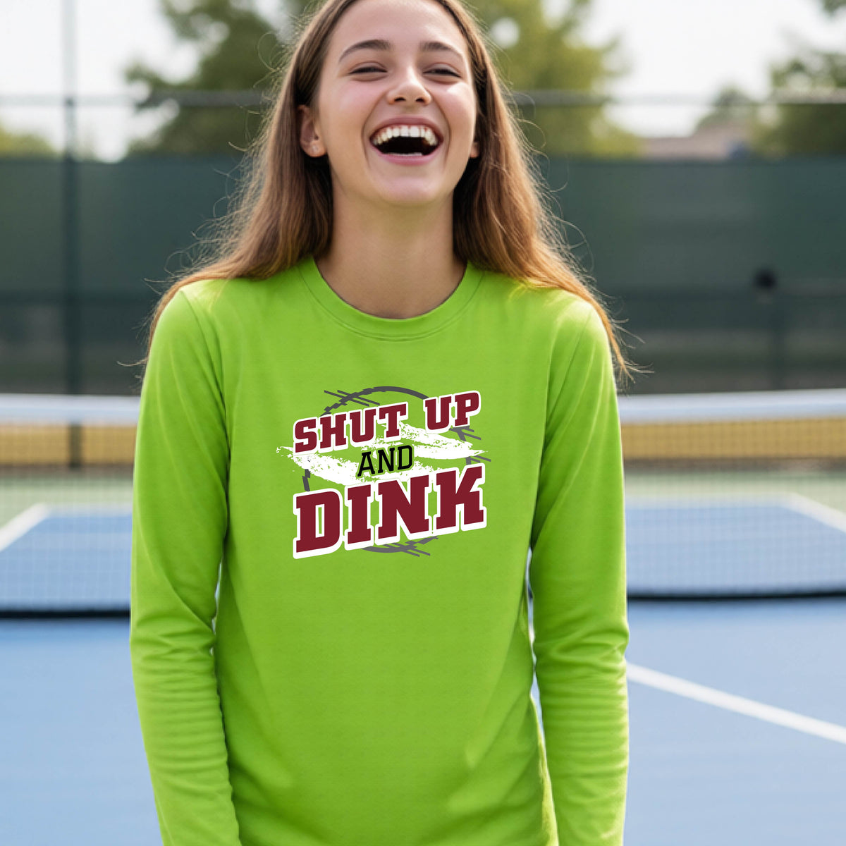 A young person on a pickleball court wearing a lime green long-sleeve t-shirt with a white, gray and black graphic of the text:  ‘Shut Up and Dink, standing on a pickleball court.