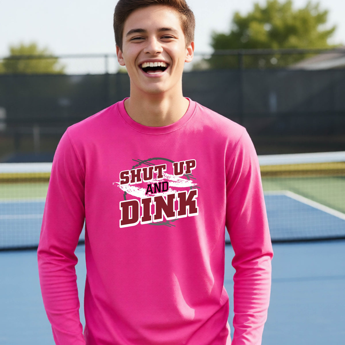 A young person on a pickleball court wearing a pink long-sleeve t-shirt with a white, gray and black graphic of the text:  ‘Shut Up and Dink, standing on a pickleball court.